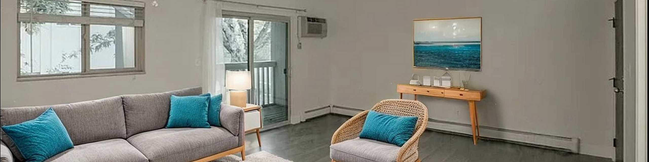 Bright and inviting living room with stylish modern furniture, featuring a cozy gray sofa with vibrant teal cushions, a wooden coffee table, and an elegant armchair. The room is enhanced by natural light through large windows and a charming
