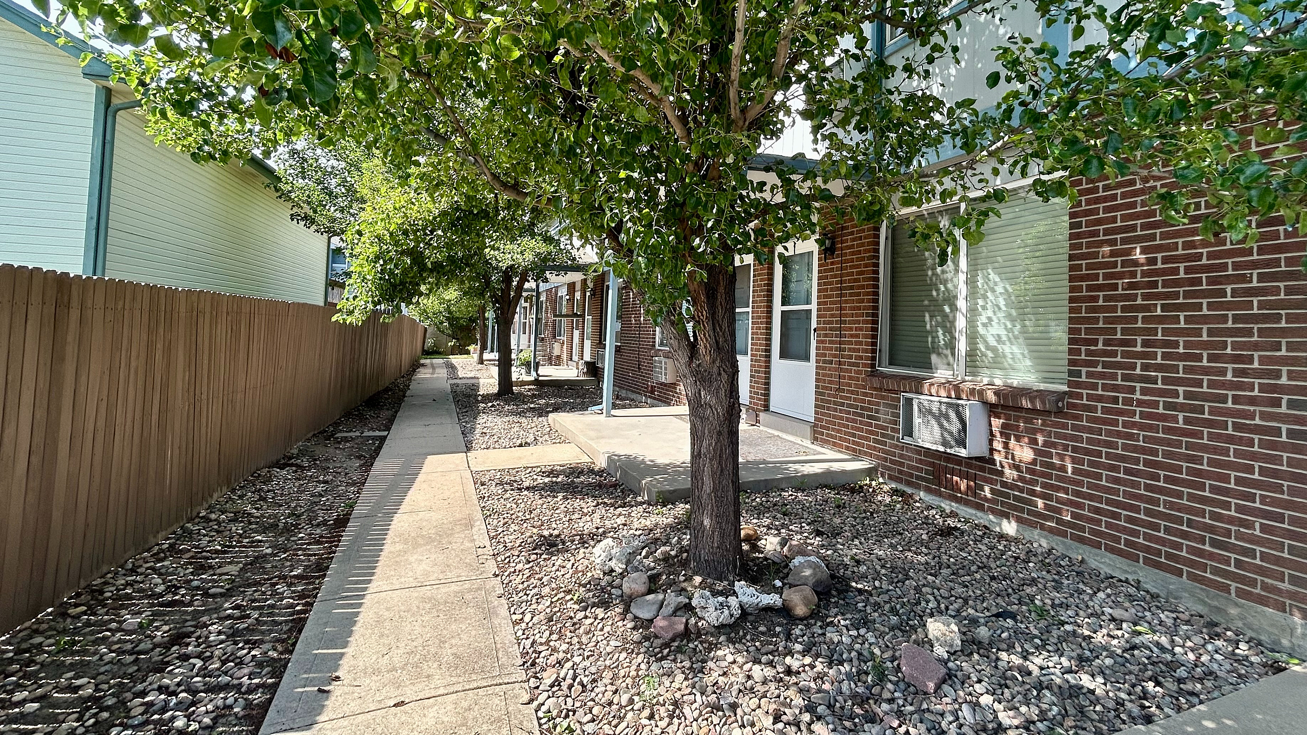 Charming apartment exterior with red brick facade, shaded walkway, and lush green trees providing a tranquil atmosphere.