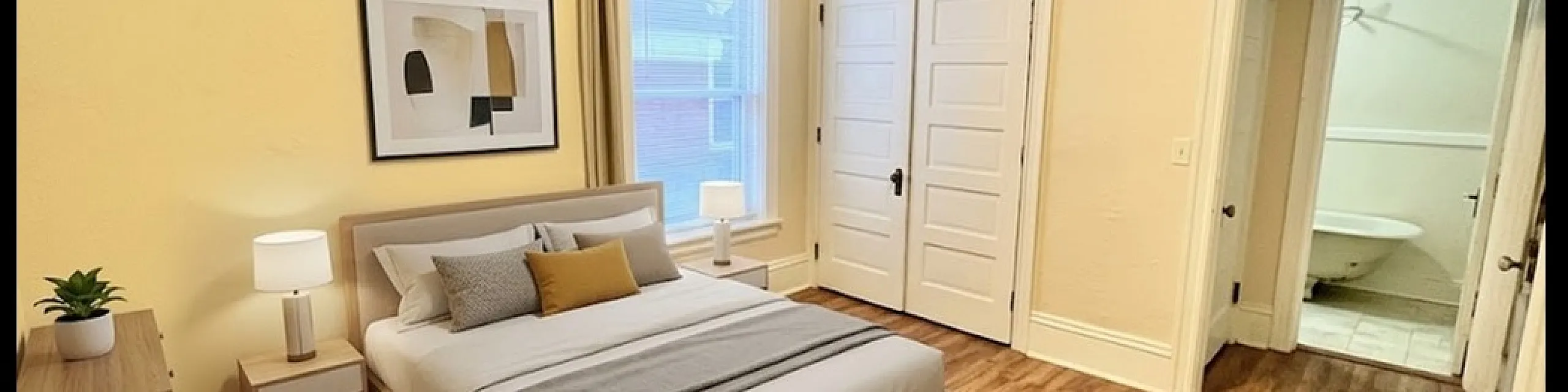 Bright and welcoming bedroom featuring elegant wood flooring, cozy bed with stylish accents, natural light from large window, and convenient access to an en-suite bathroom.
