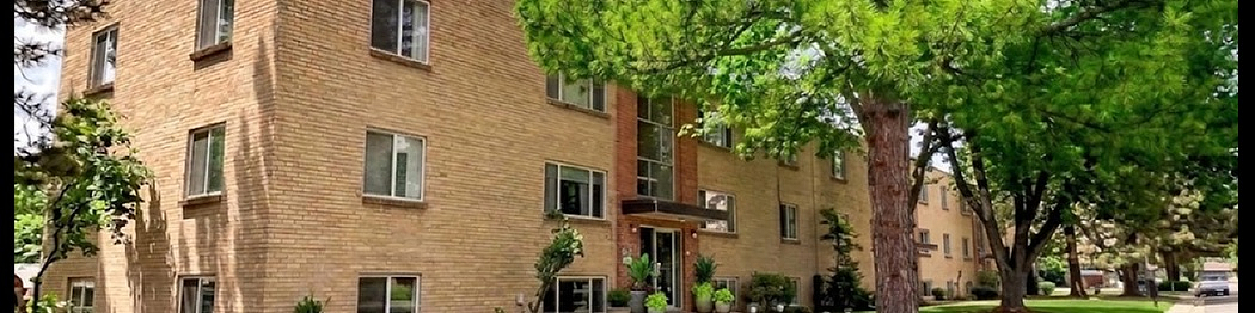Charming brick apartment building with lush green landscaping, set in a serene neighborhood with beautiful mature trees.