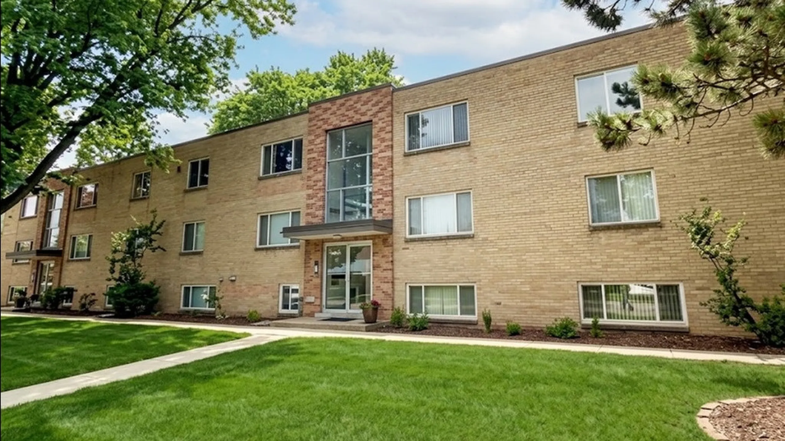 Charming brick apartment building surrounded by lush green grass and mature trees, highlighting a modern living entrance and ample windows for natural light.