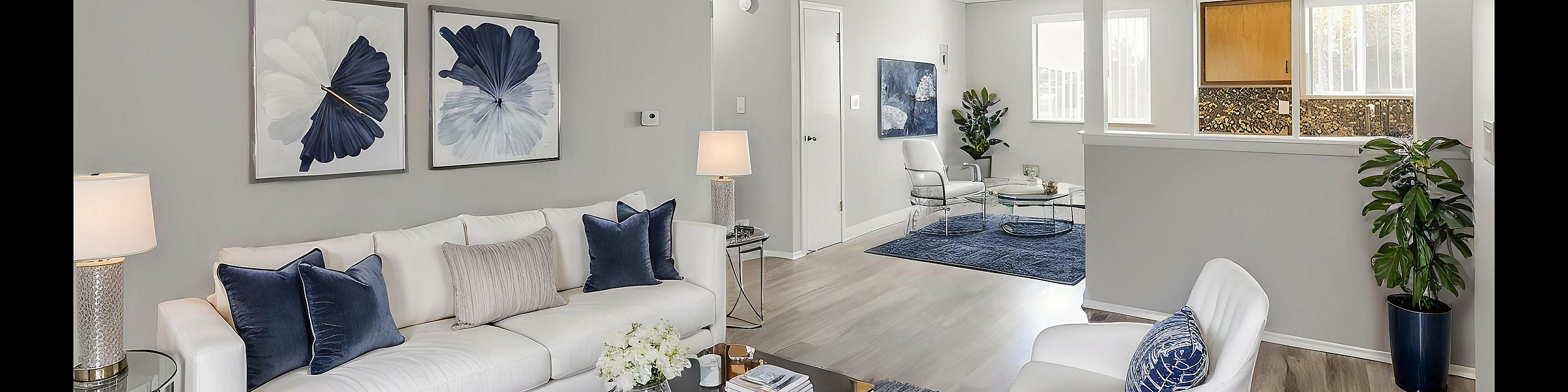 Bright and modern living room with elegant decor, plush white sofa, blue accents, and open layout leading to a cozy seating area and stylish kitchen.