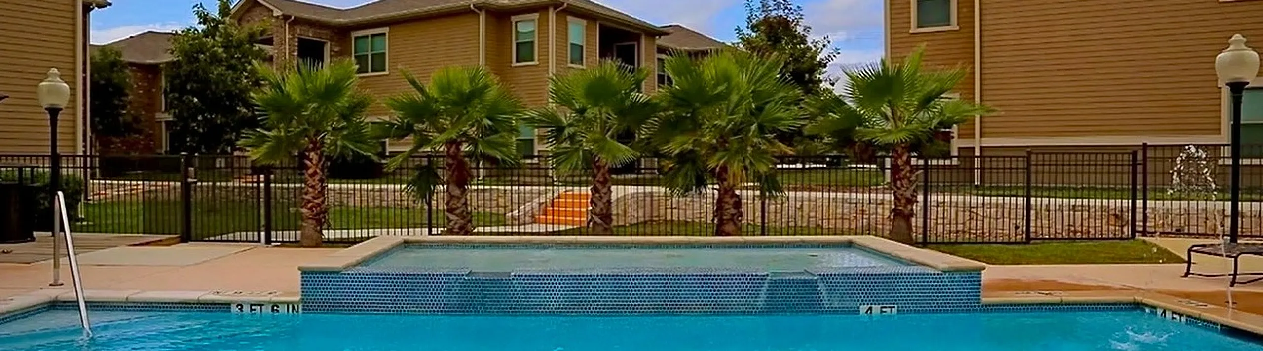 Inviting outdoor pool surrounded by lush palm trees, set against elegant two-story apartment buildings and a bright blue sky.