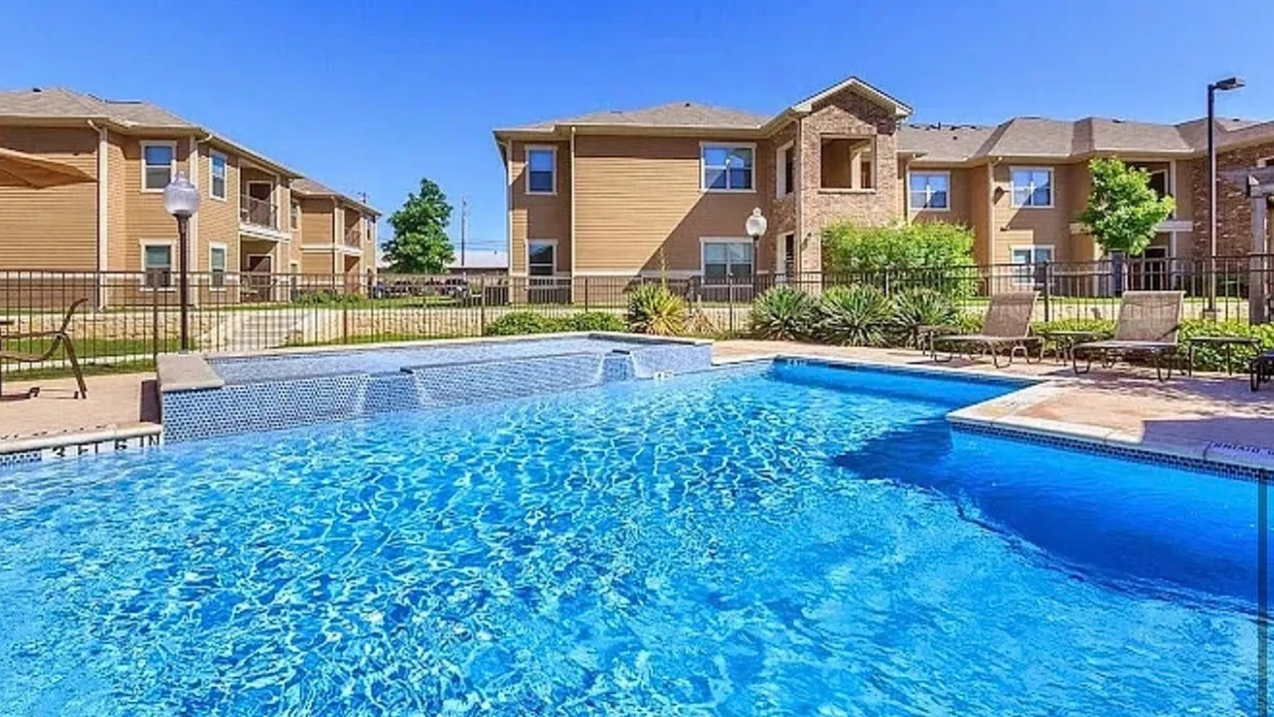 Inviting apartment complex with a crystal-clear swimming pool, surrounded by sun loungers and beautifully landscaped grounds under a bright blue sky.
