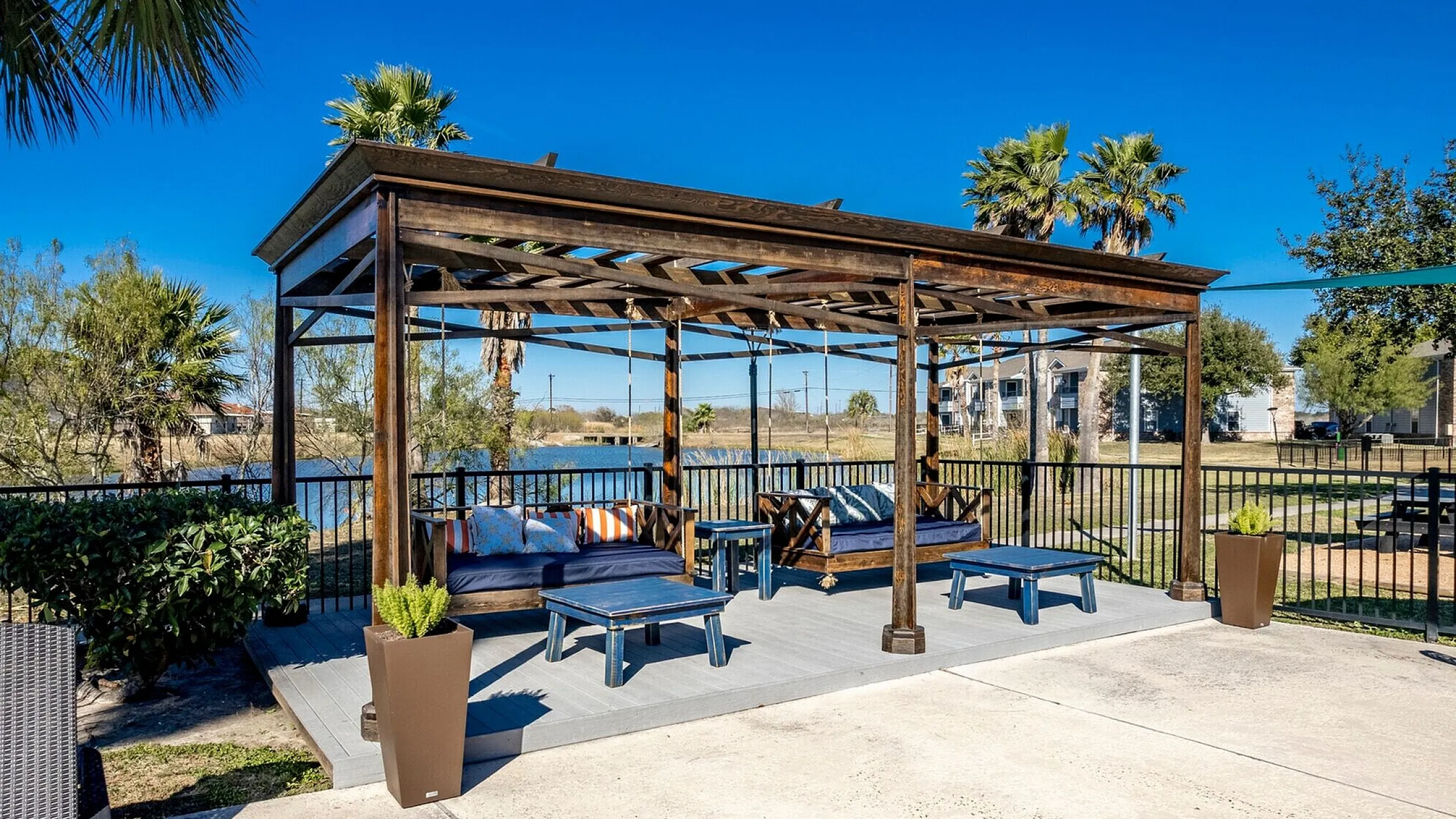 Charming outdoor seating area with a wooden pergola, cozy sofas, and a picturesque view of palm trees and a serene lake.