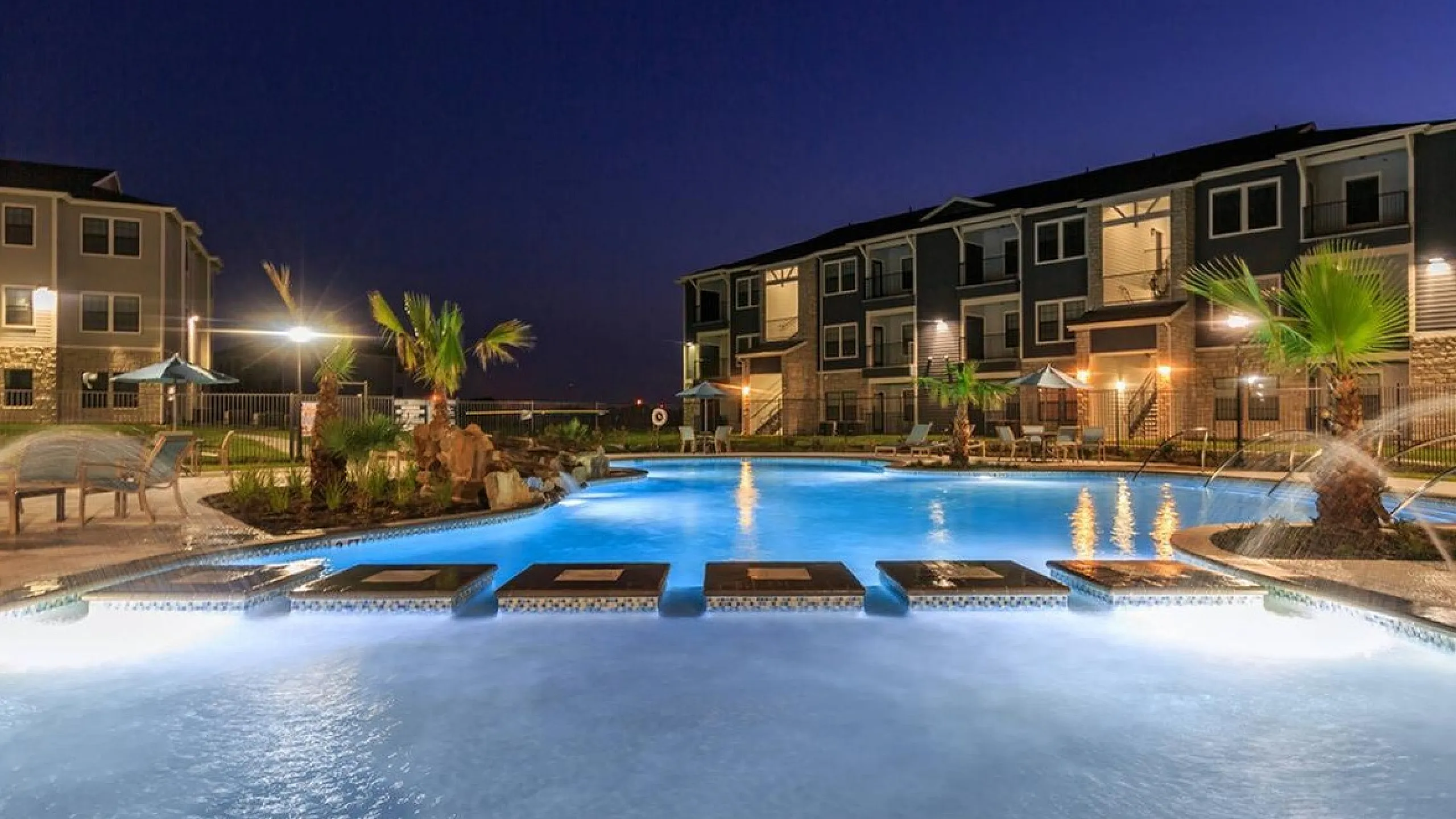 Luxurious apartment complex at night showcasing an illuminated swimming pool surrounded by palm trees and modern architecture.