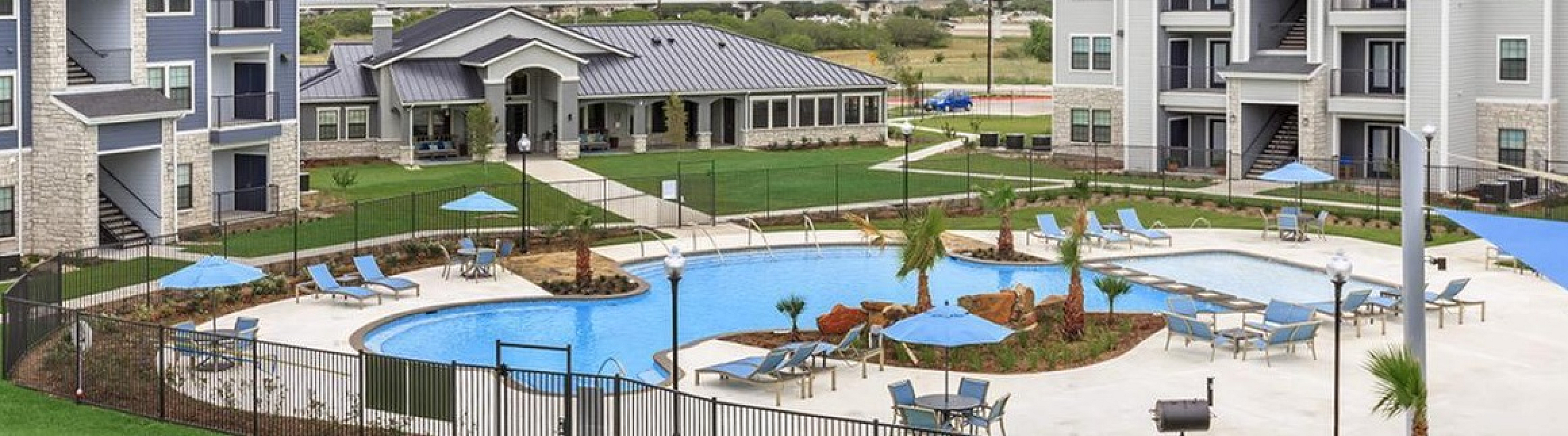 Charming apartment complex with modern buildings, lush green lawns, and a sparkling swimming pool surrounded by comfortable lounge chairs.