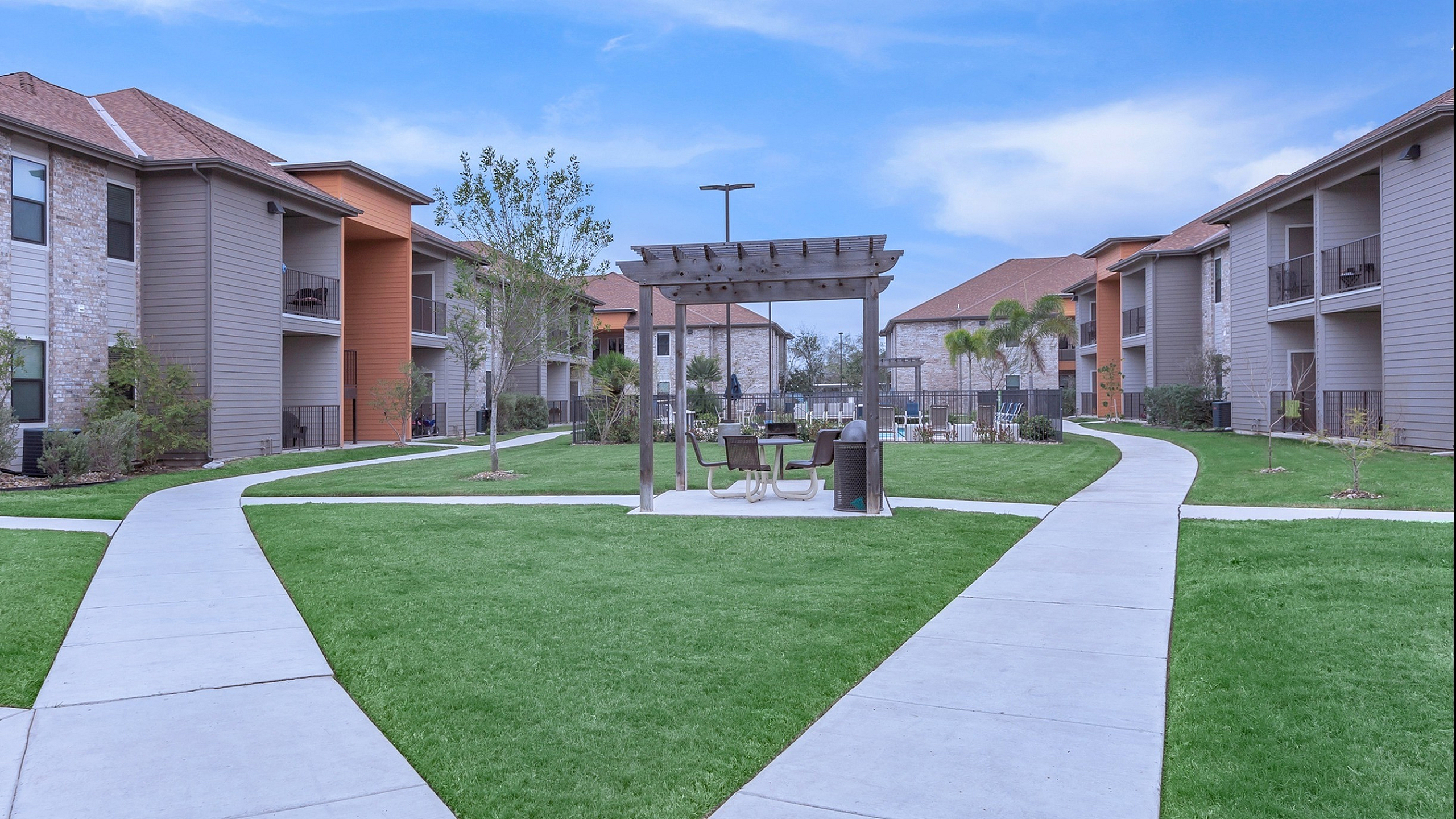 Charming apartment complex courtyard featuring immaculate green lawns, modern architecture, and a cozy pergola seating area.