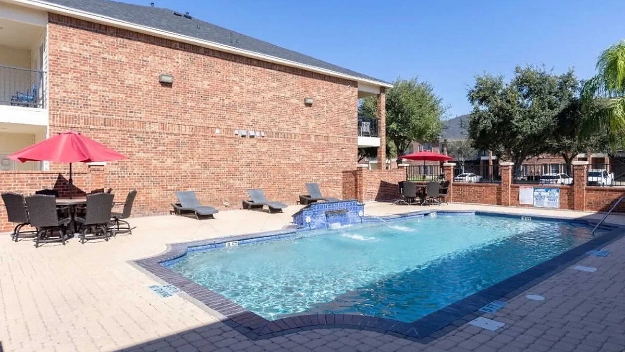 Inviting outdoor pool area with elegant brick surroundings, relaxing lounge chairs, and vibrant red umbrellas offering shade and style.