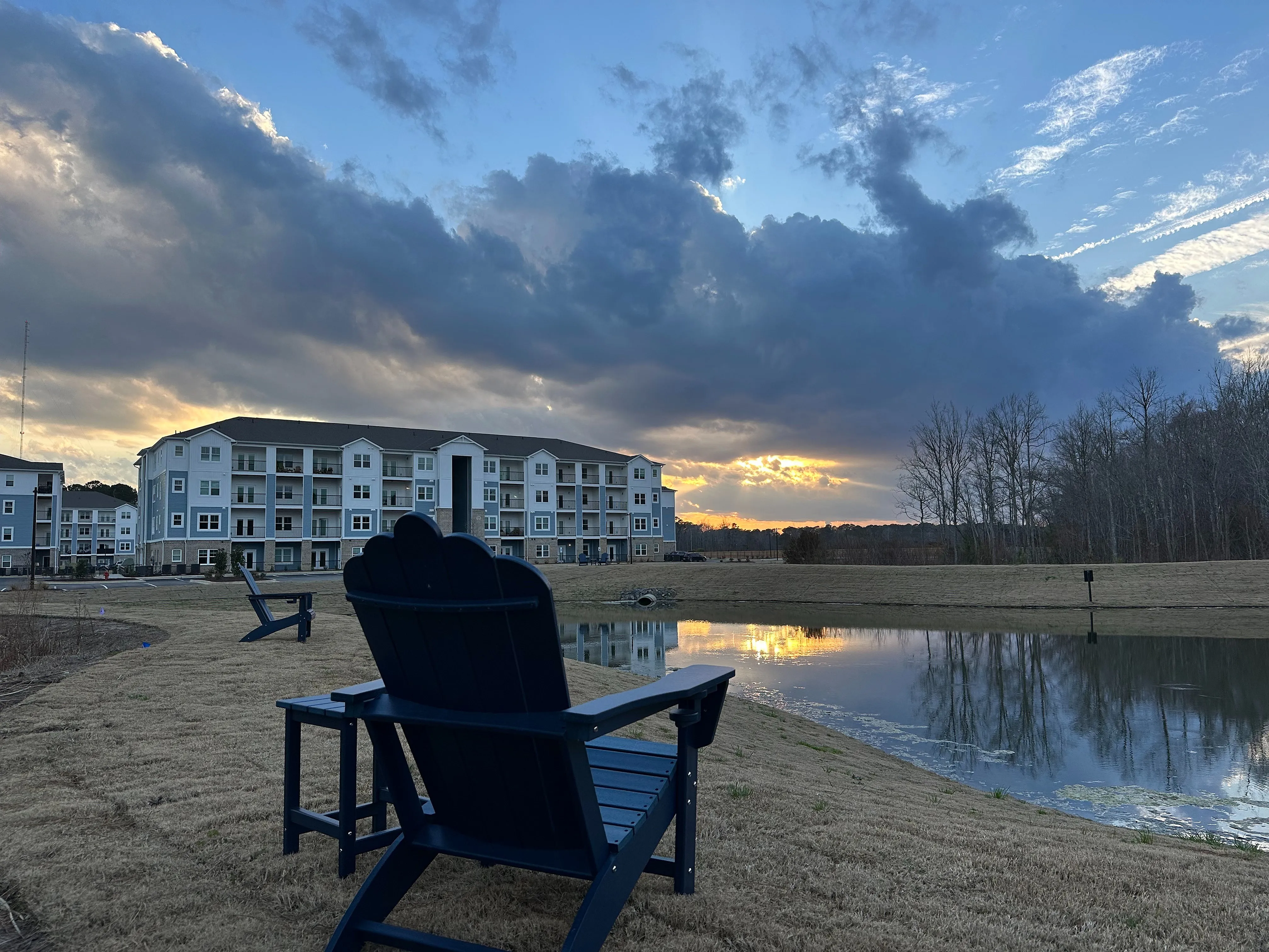 Charming lakeside apartment complex with Adirondack chairs, reflecting a picturesque sunset sky.