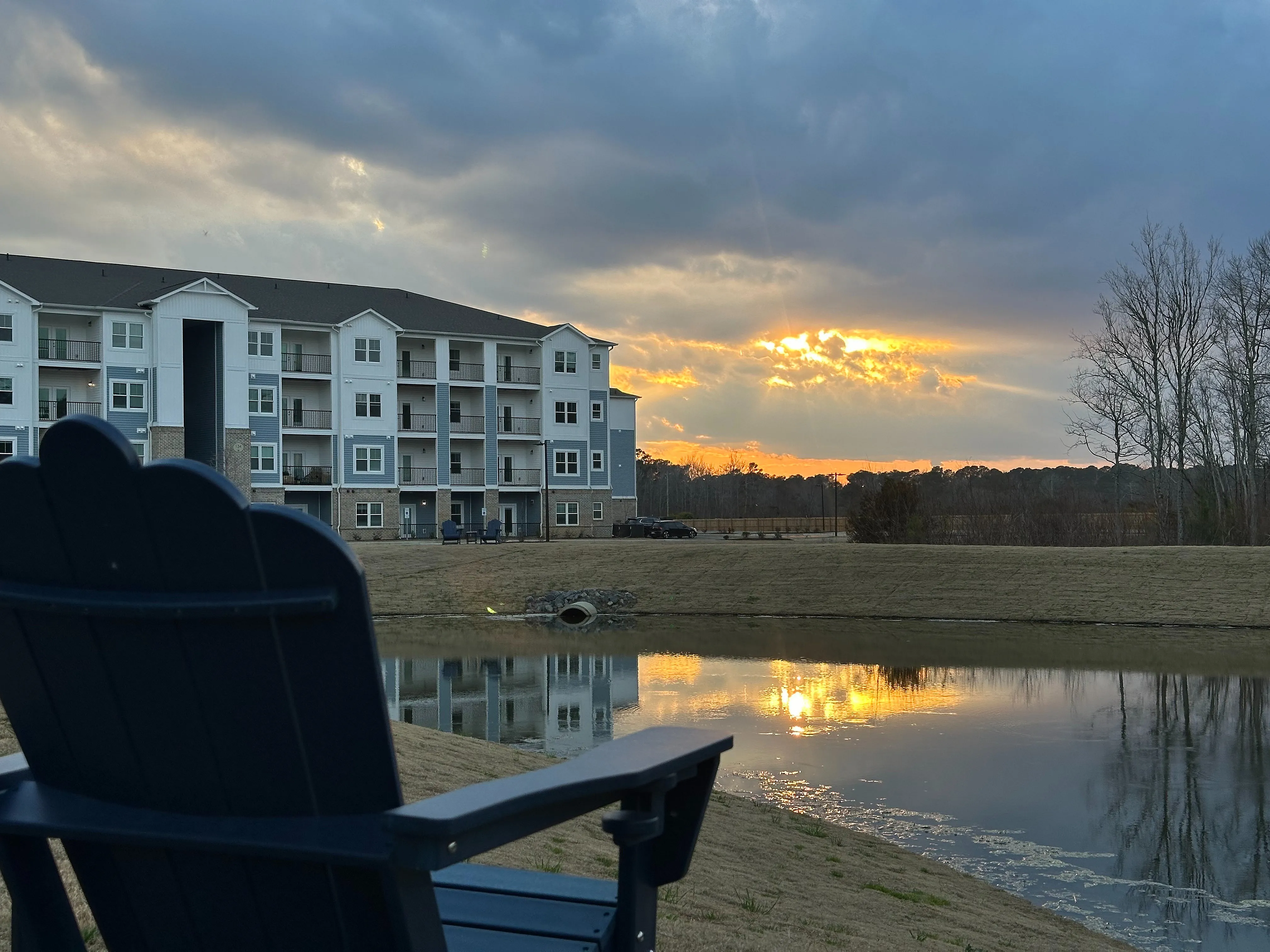 Charming apartment complex by a tranquil pond at sunset, with a scenic view and comfortable outdoor seating.