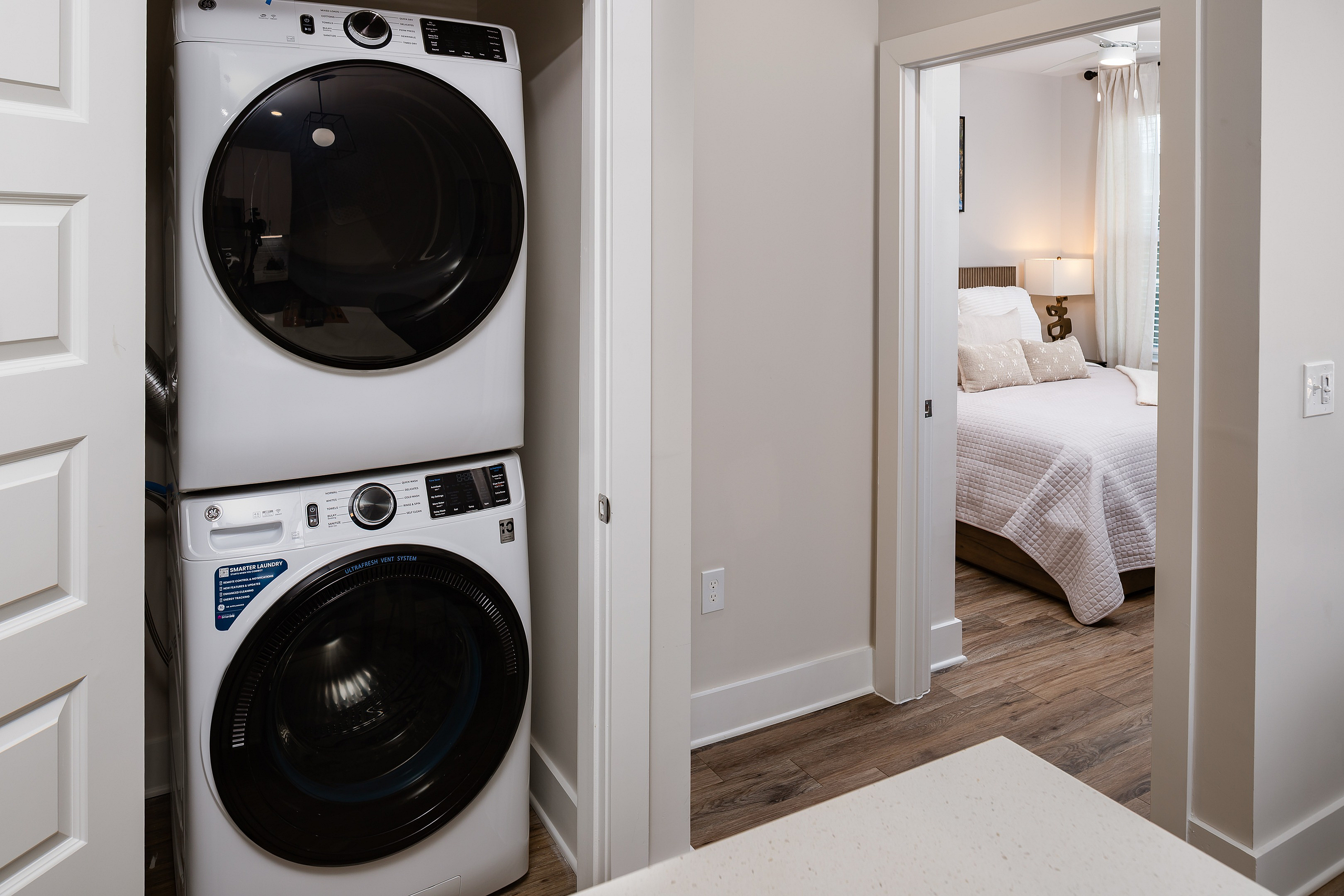 Chic apartment feature with modern stacked washer and dryer next to a cozy bedroom illuminated by natural light.