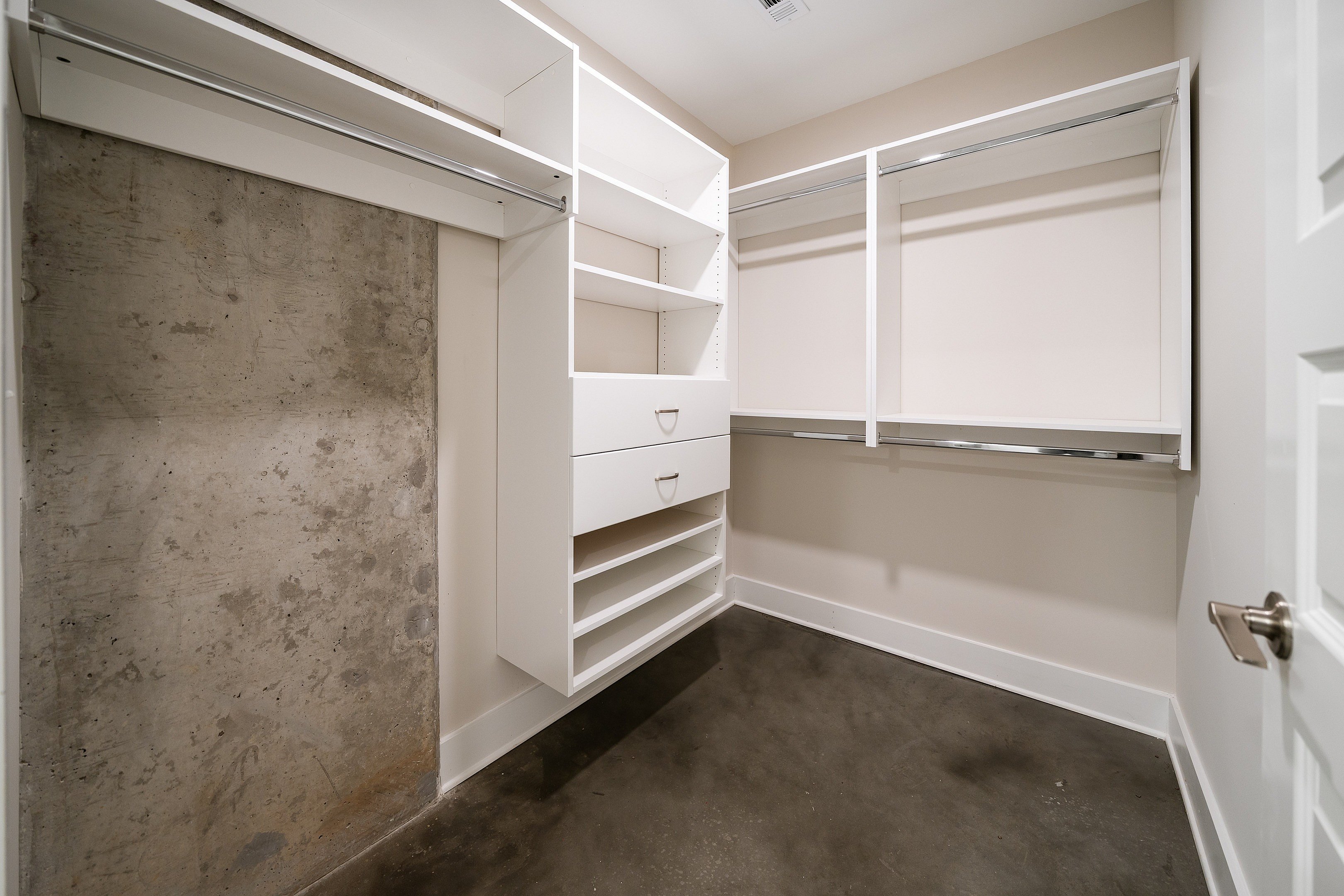 Modern walk-in closet with sleek white shelves, drawers, and ample hanging space against an industrial concrete wall.