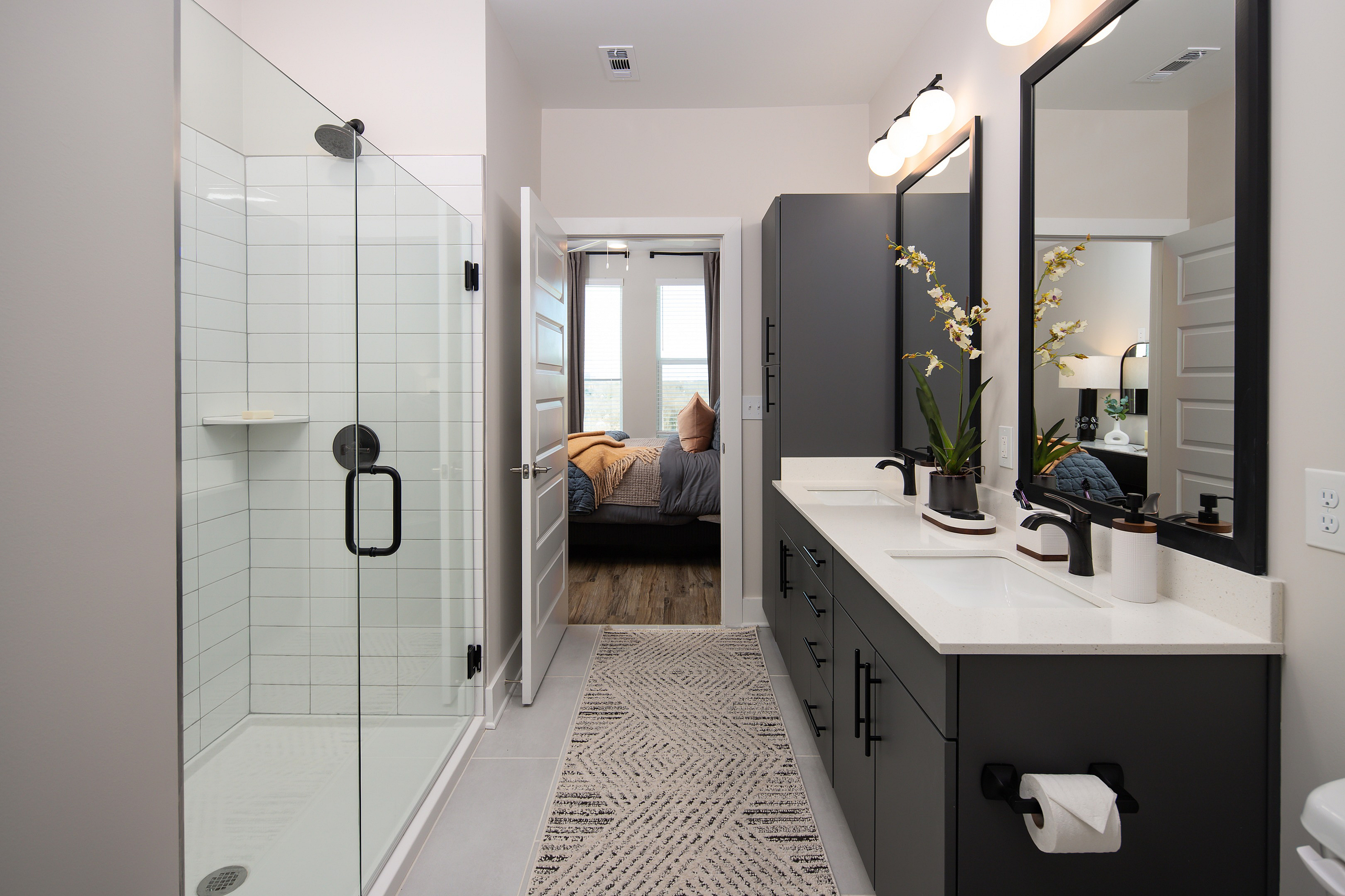 Elegant modern bathroom with sleek double vanity, spacious glass shower, and view into a cozy bedroom.
