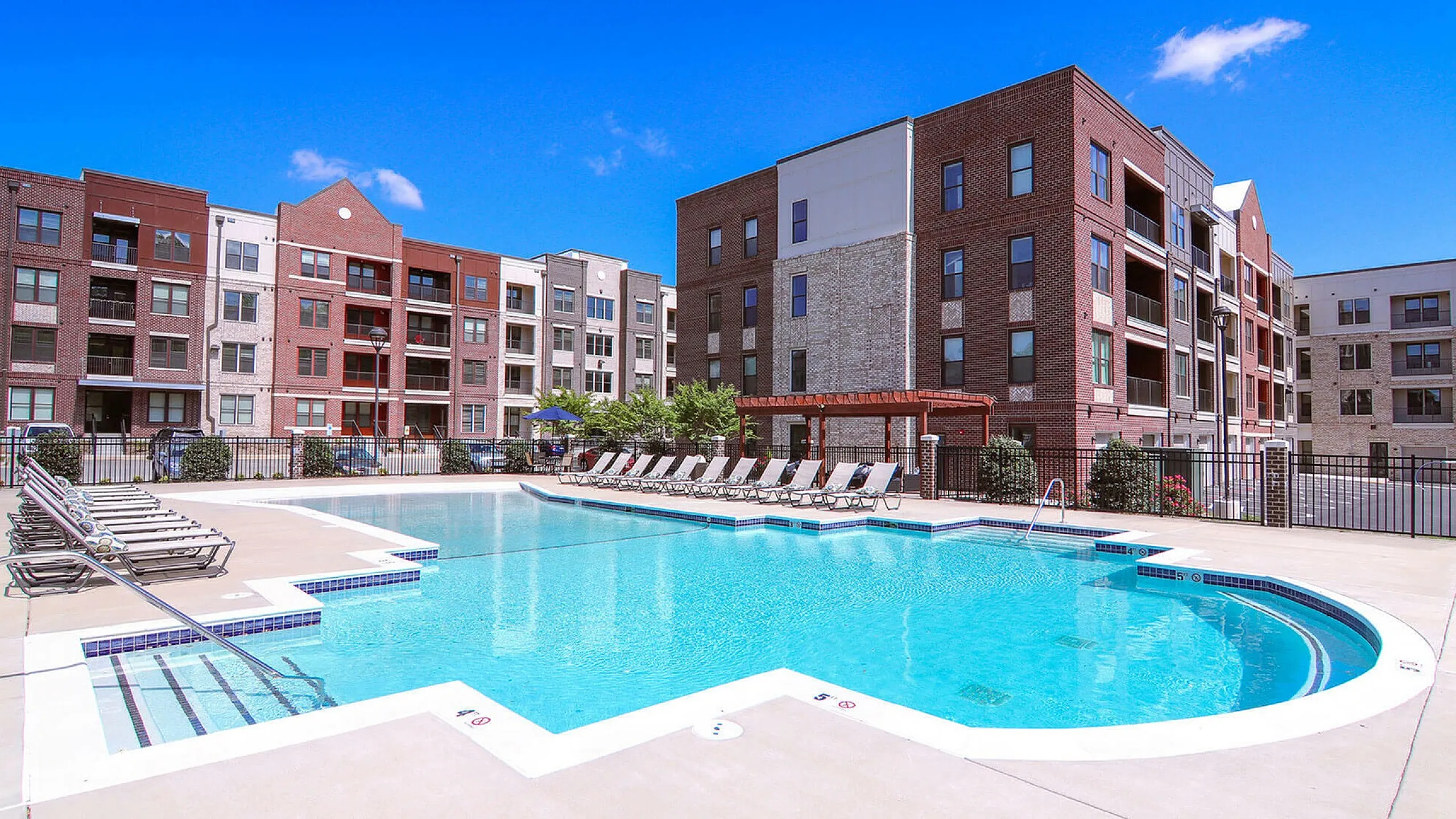 Modern apartment complex with a sparkling outdoor pool and ample lounge chairs under a clear blue sky.
