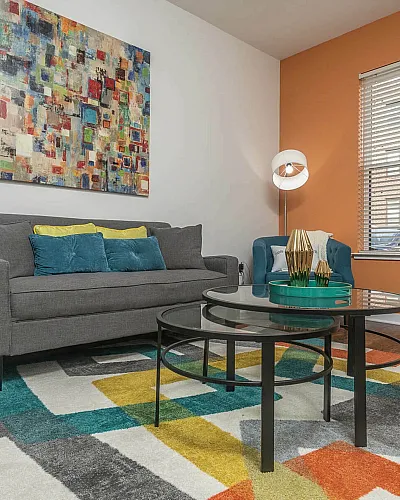 Modern living room with a gray sofa, colorful abstract art, geometric rug, and large windows.