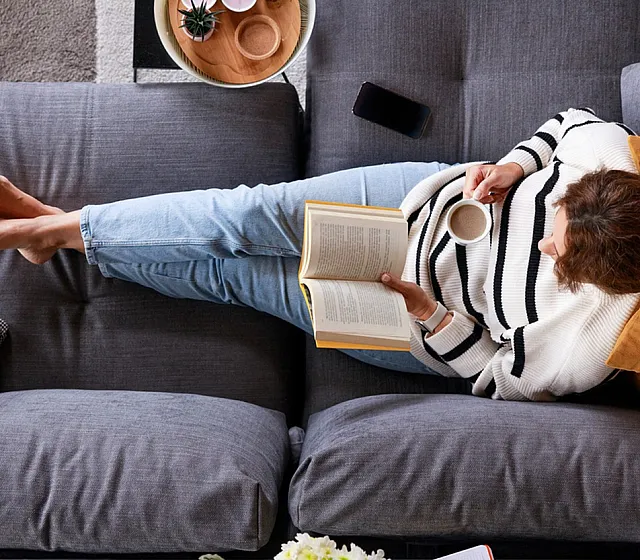 Comfortable living room setup with a person relaxing on a gray sofa, enjoying a book and coffee.