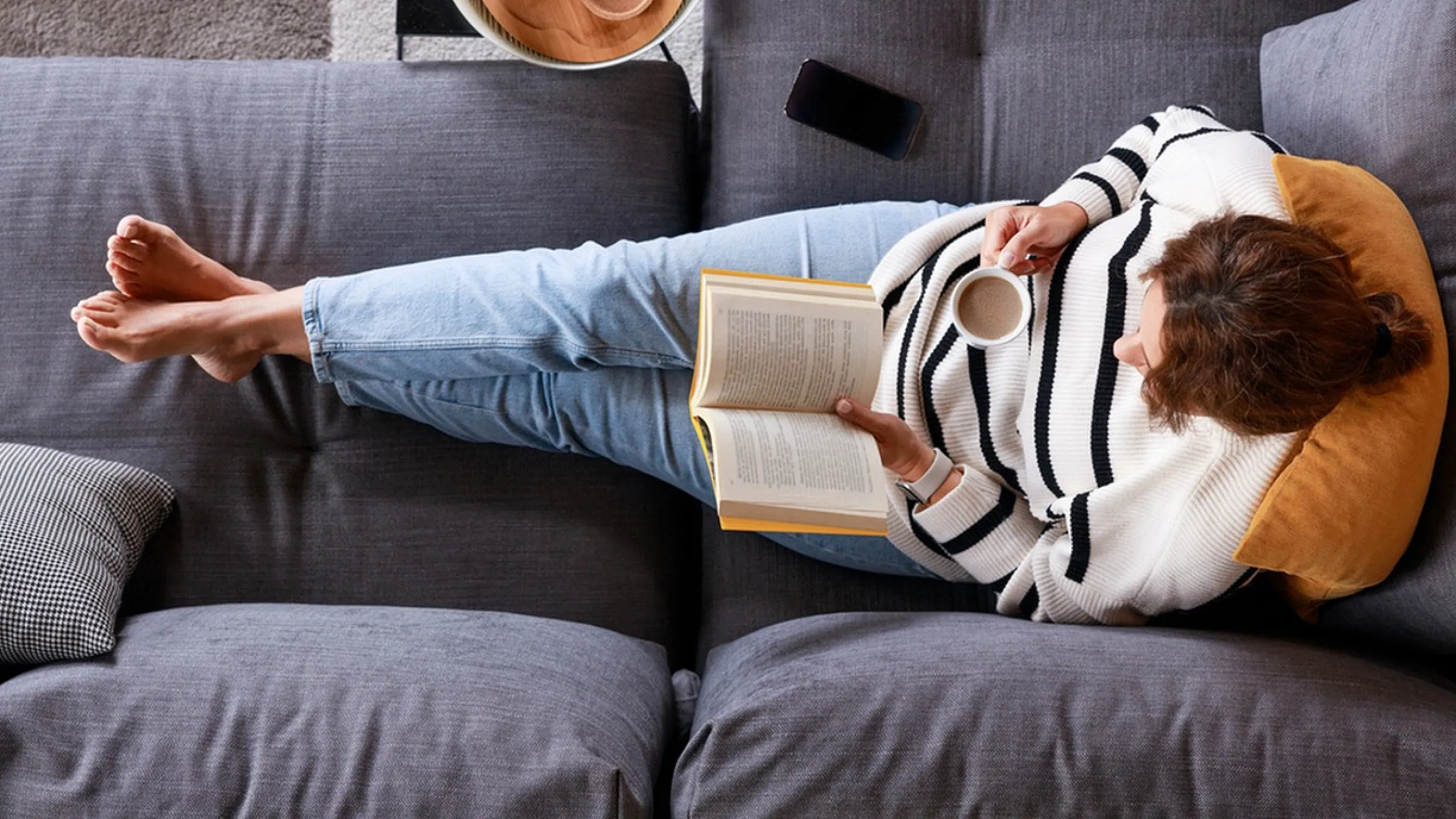 Top view of a person relaxing on a stylish grey sofa with a book and coffee in a modern living room setting.