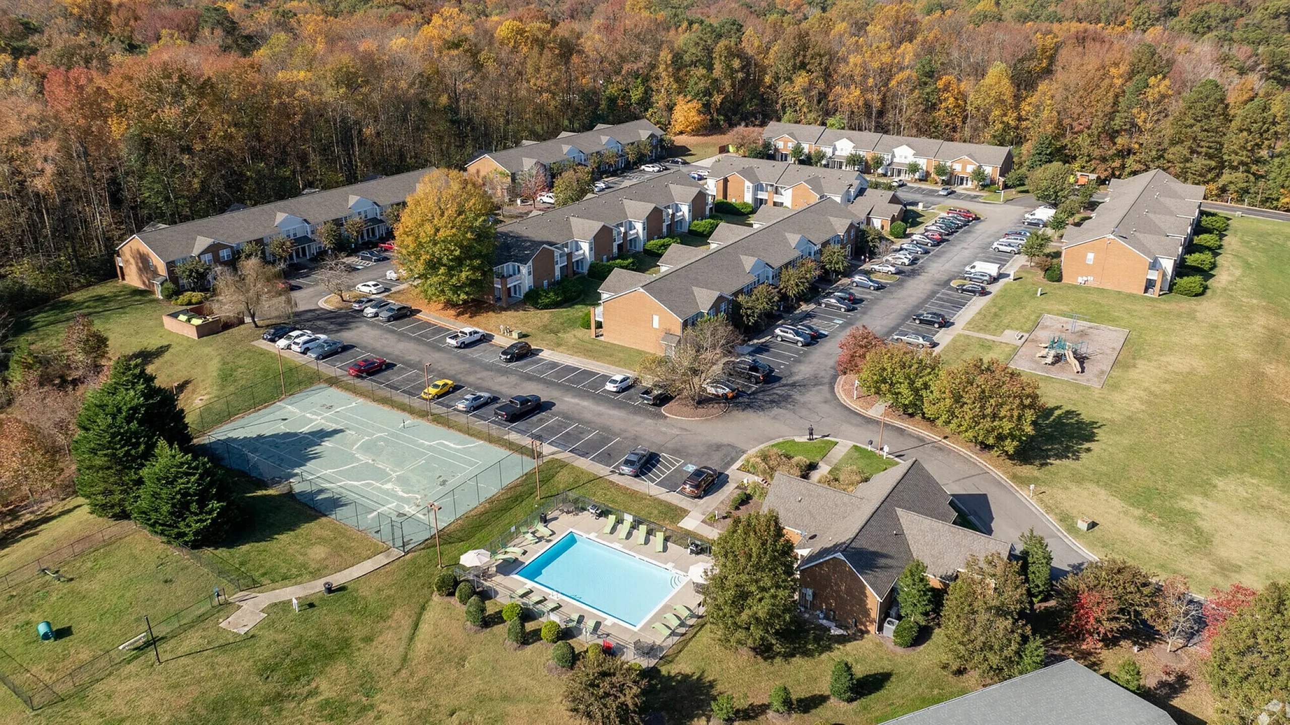Aerial view of a serene apartment complex with a pool, tennis court, and lush autumn foliage.