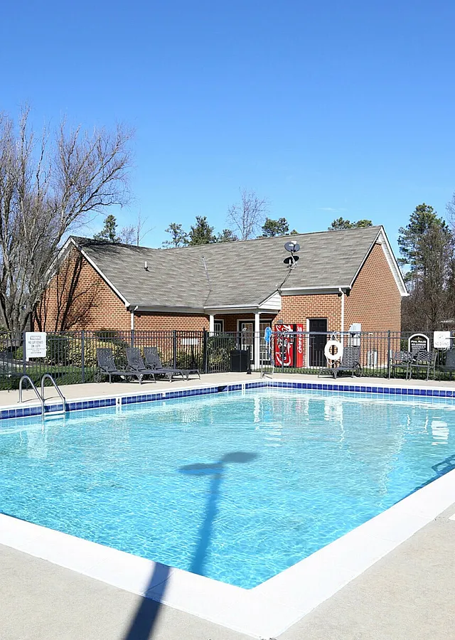 Sparkling outdoor swimming pool surrounded by lounge chairs, with a brick clubhouse in the background.