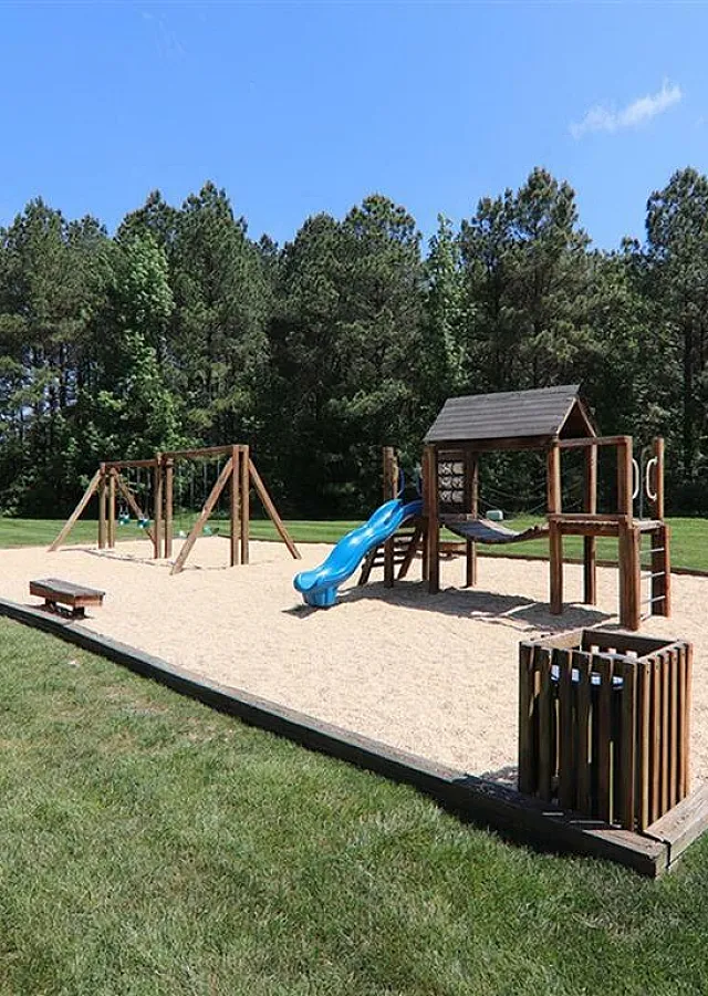 Beautiful playground with swings, slide, and climbing structure set within a spacious grassy area and surrounded by lush trees.