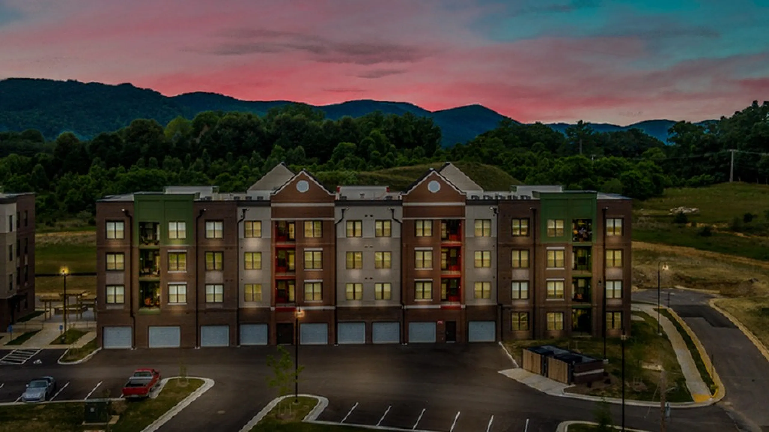 Evening view of a modern apartment complex with illuminated windows and scenic mountain backdrop.