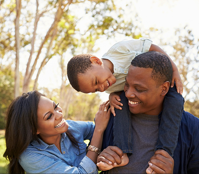 A joyful family enjoying a sunny day in the park, with a young child playfully sitting on a parent's shoulders.
