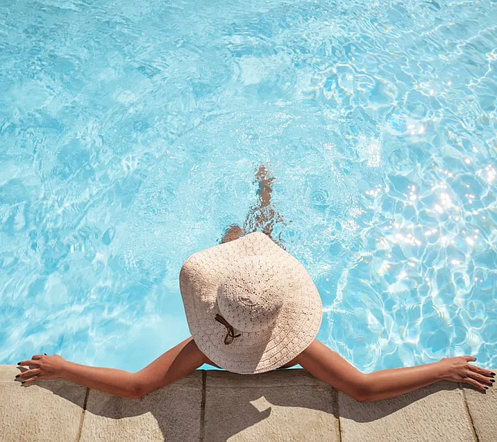 A person relaxing at the edge of a swimming pool wearing a sun hat, with clear blue water reflecting sunlight.