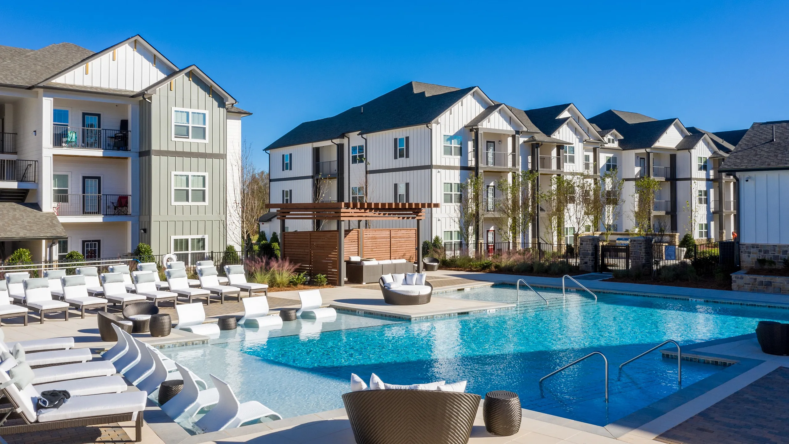 Modern apartment complex with a spacious outdoor pool area, surrounded by lounge chairs and lush greenery.