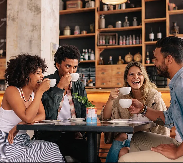Group of friends enjoying coffee and laughter in a cozy caf&eacute; setting with rustic decor.