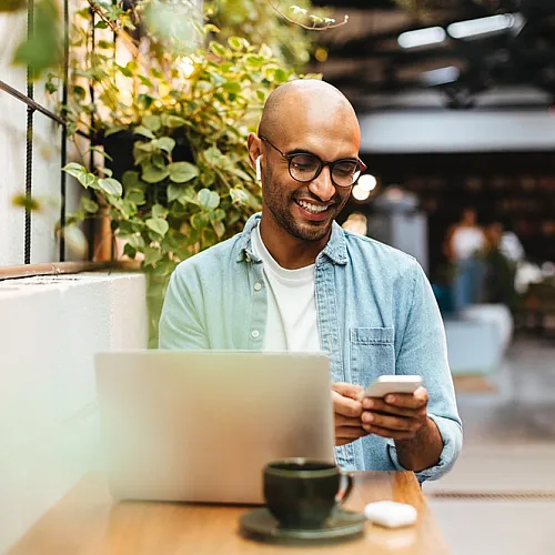 Man smiling while using a smartphone in a modern cafe with a laptop and green plants.