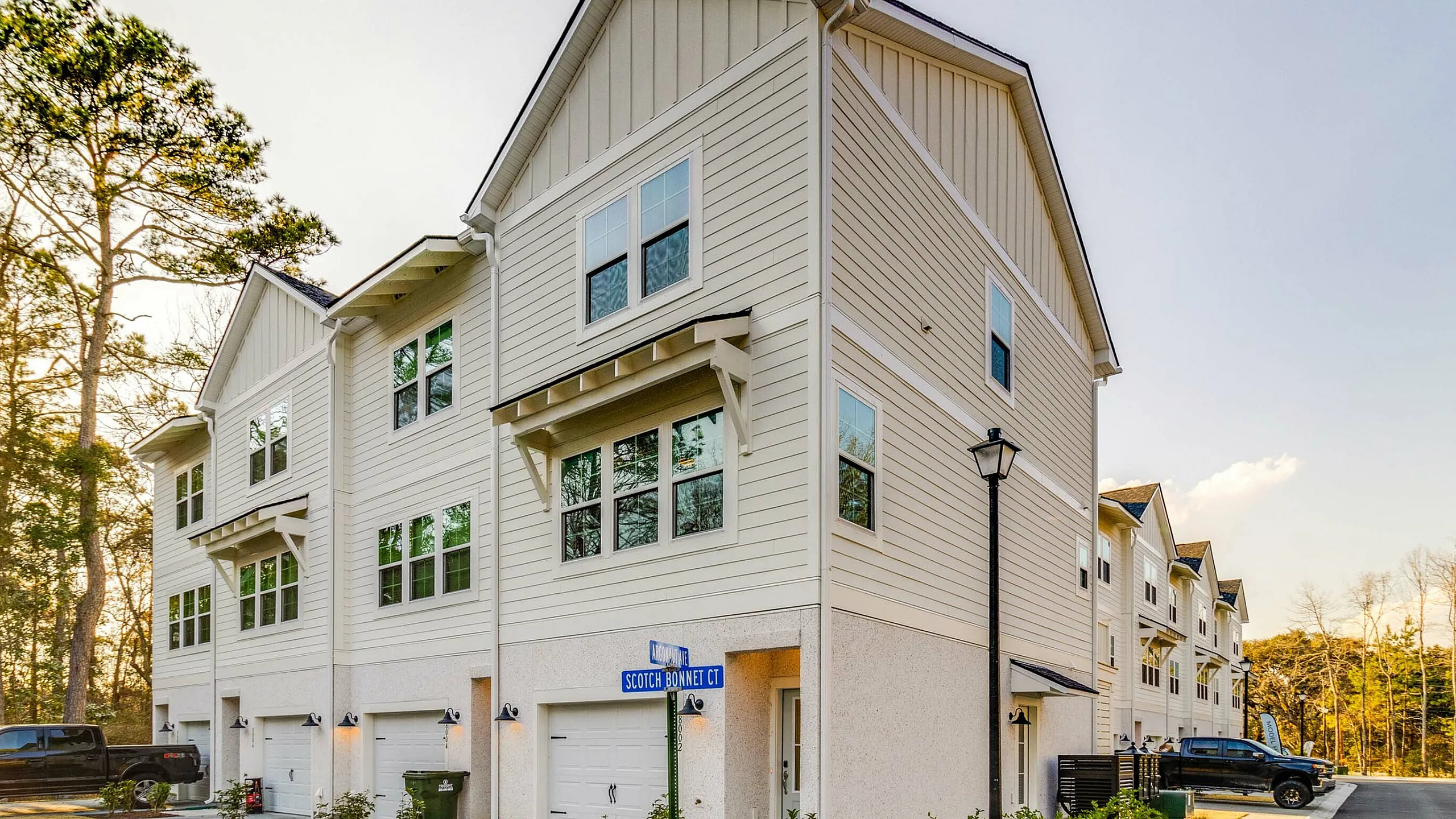 Contemporary multi-story townhouse with elegant siding, large windows, and spacious garages, set on a serene tree-lined street.