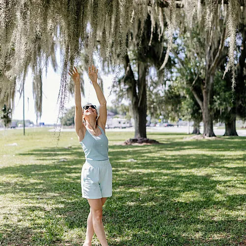 Joyful woman reaching for Spanish moss under a sunlit tree, enjoying a peaceful day outdoors.