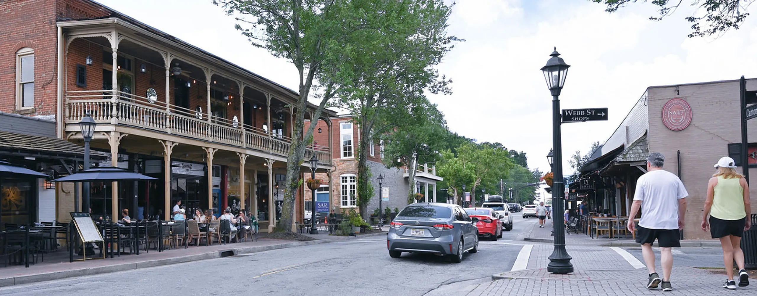 Charming downtown street scene showcasing historic brick buildings, bustling outdoor cafes, and lush green trees under a bright blue sky.