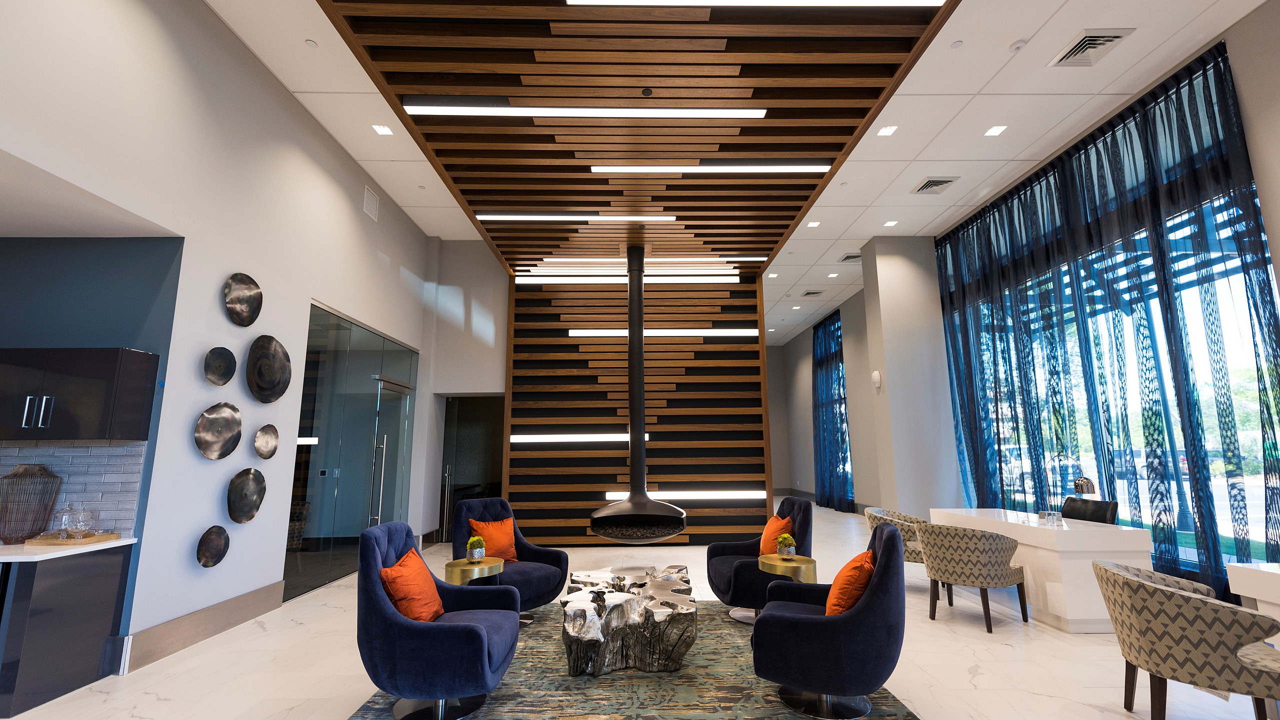 Lobby of an apartment building featuring mid-century modern design, with warm wood paneling, geometric light fixtures, minimalist furniture, and large windows letting in natural light.
