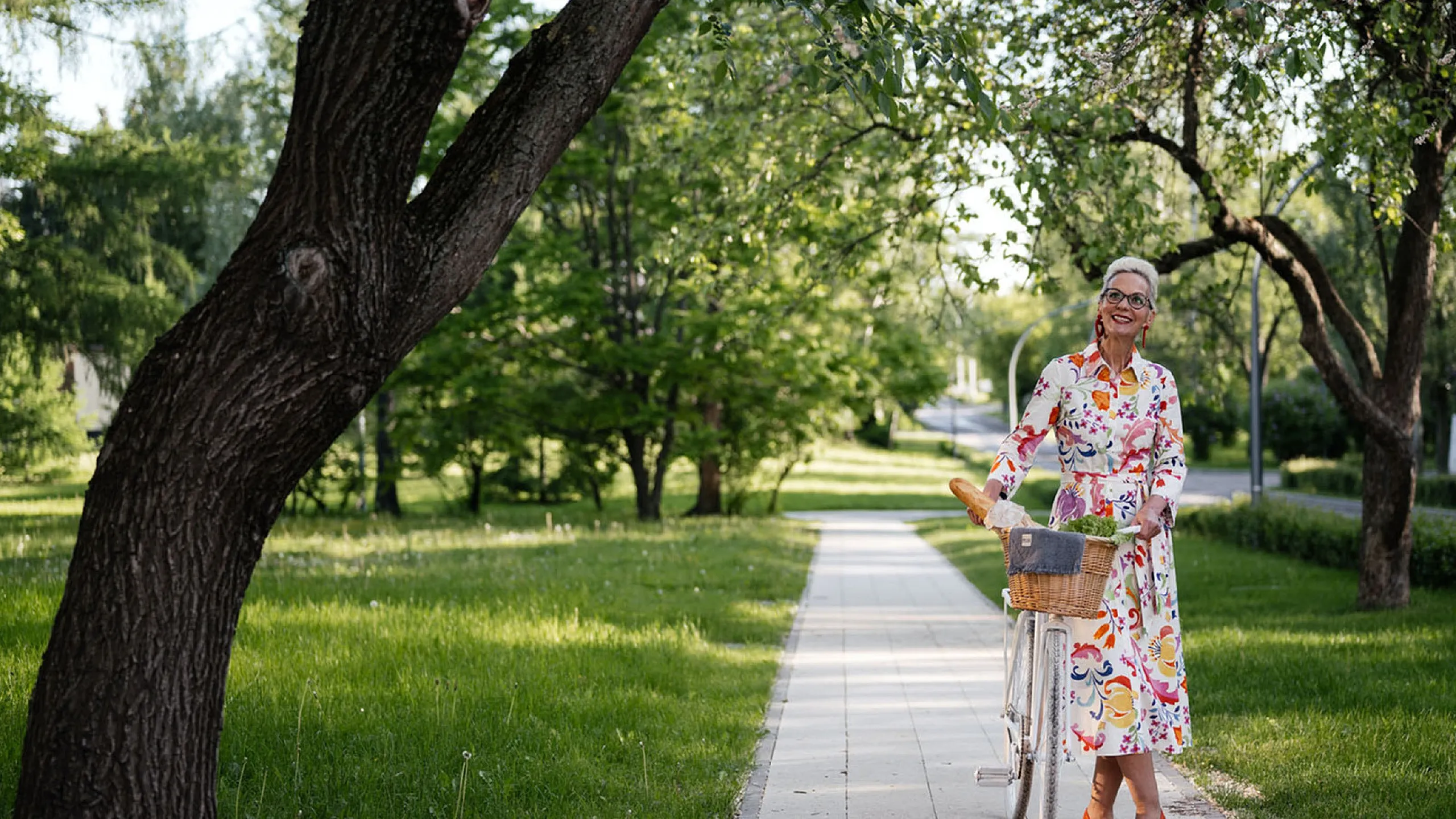 Charming woman in vibrant floral dress enjoying a peaceful park stroll with a classic bicycle, surrounded by lush green scenery and blooming trees.