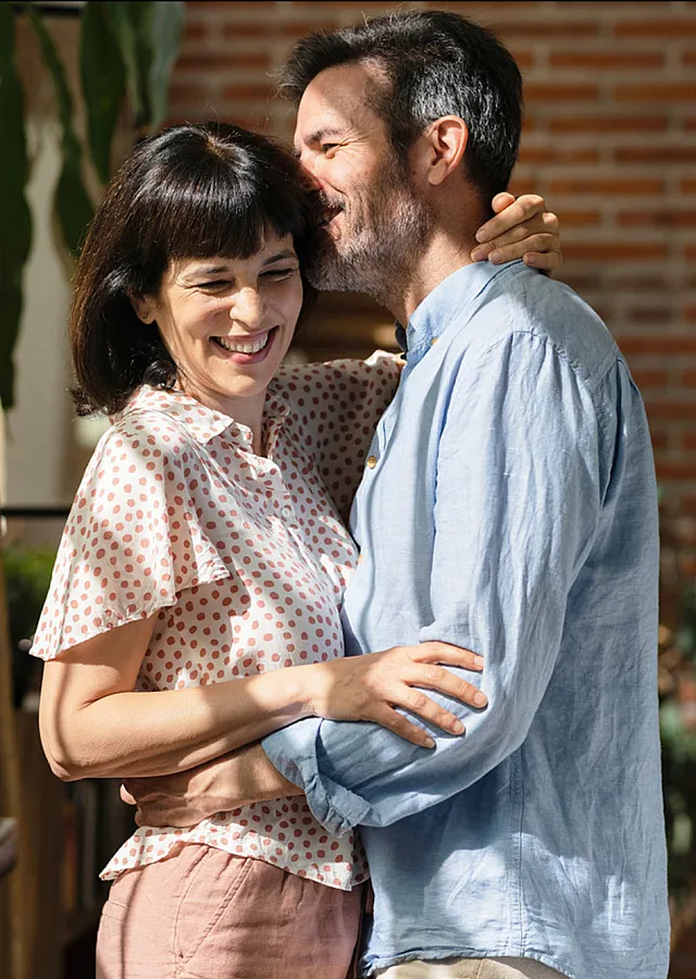 Cheerful couple embracing in a sunlit, stylish living room with exposed brick walls and lush greenery, showcasing a cozy and inviting apartment atmosphere.