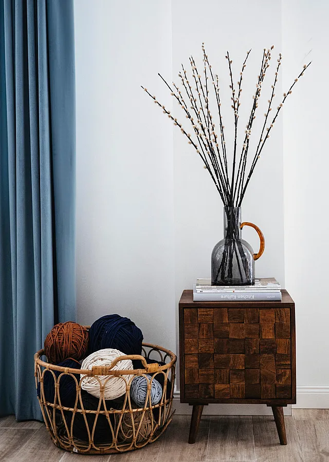Chic corner decor with textured wooden cabinet, elegant vase arrangement, and cozy yarn basket beside soft blue drapes.