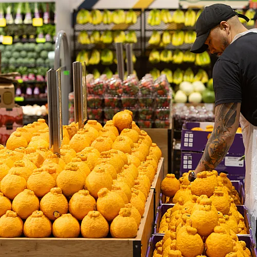 Vibrant grocery store display featuring a colorful stack of fresh yellow-orange fruits meticulously arranged, with a dedicated staff member ensuring quality presentation.