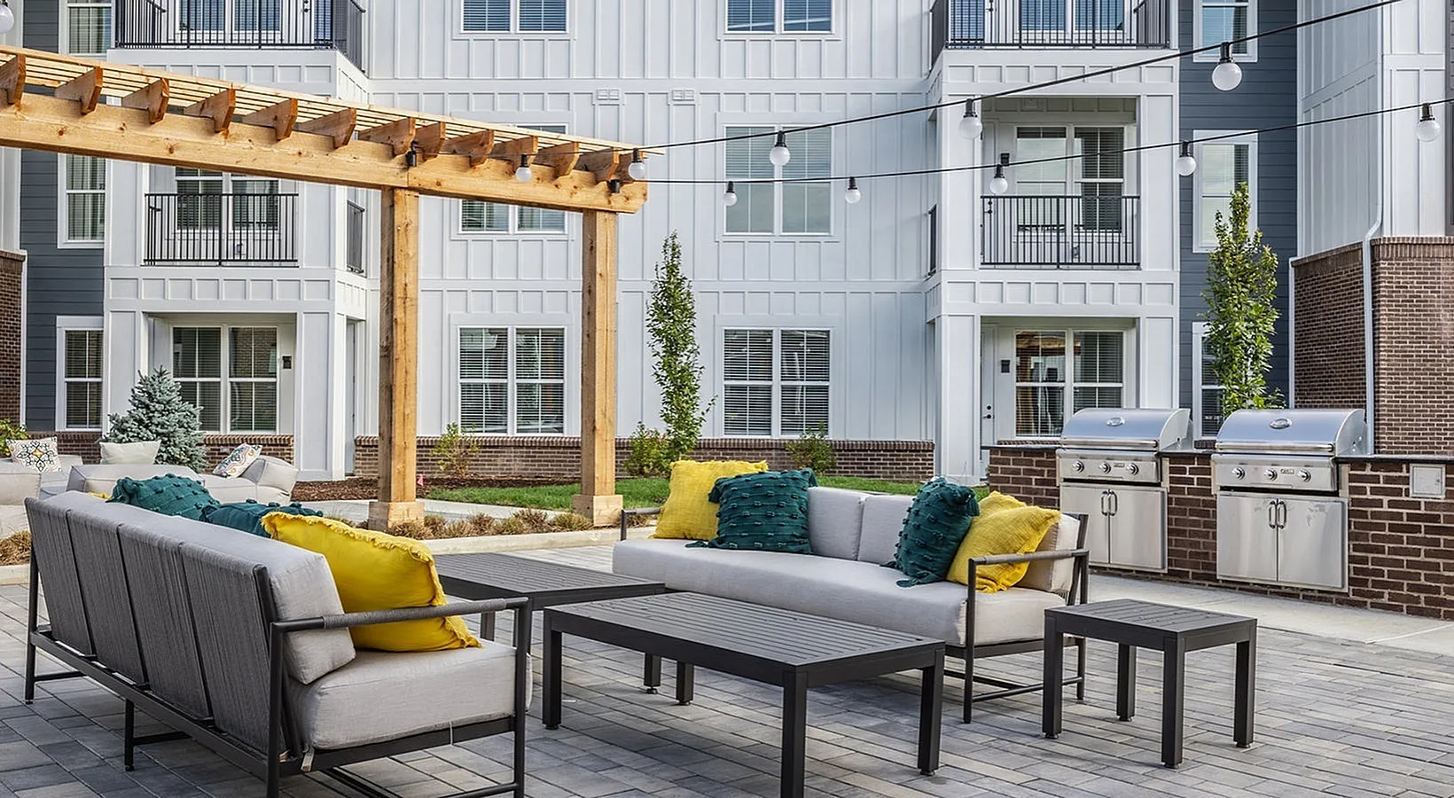 Chic outdoor patio with modern seating under a wooden pergola, featuring vibrant cushions, string lights, and stainless grills against a stylish apartment backdrop.