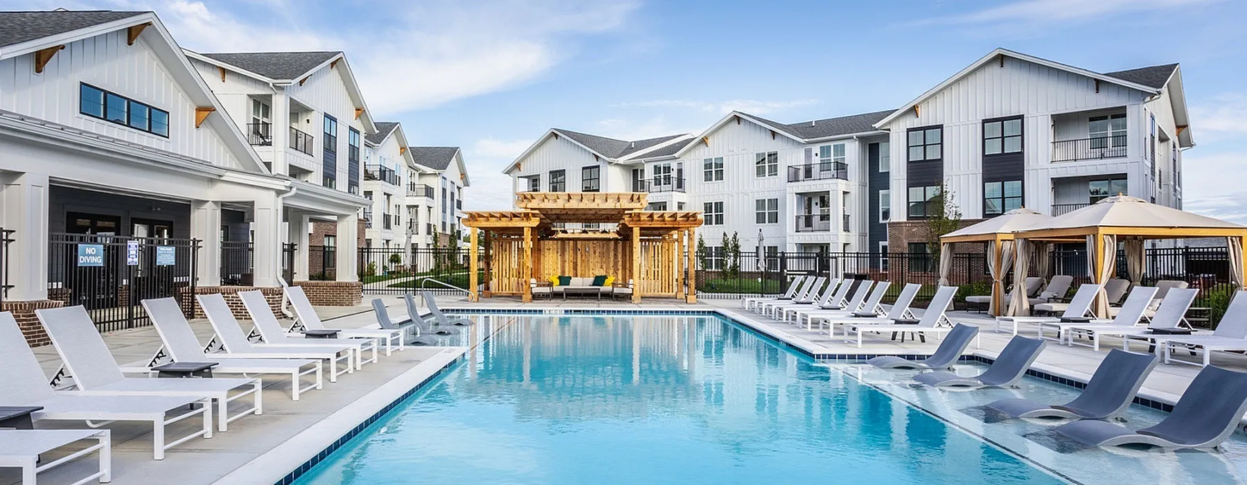 Luxurious apartment complex featuring a sparkling swimming pool, modern lounge chairs, and inviting wooden cabana under a clear blue sky.