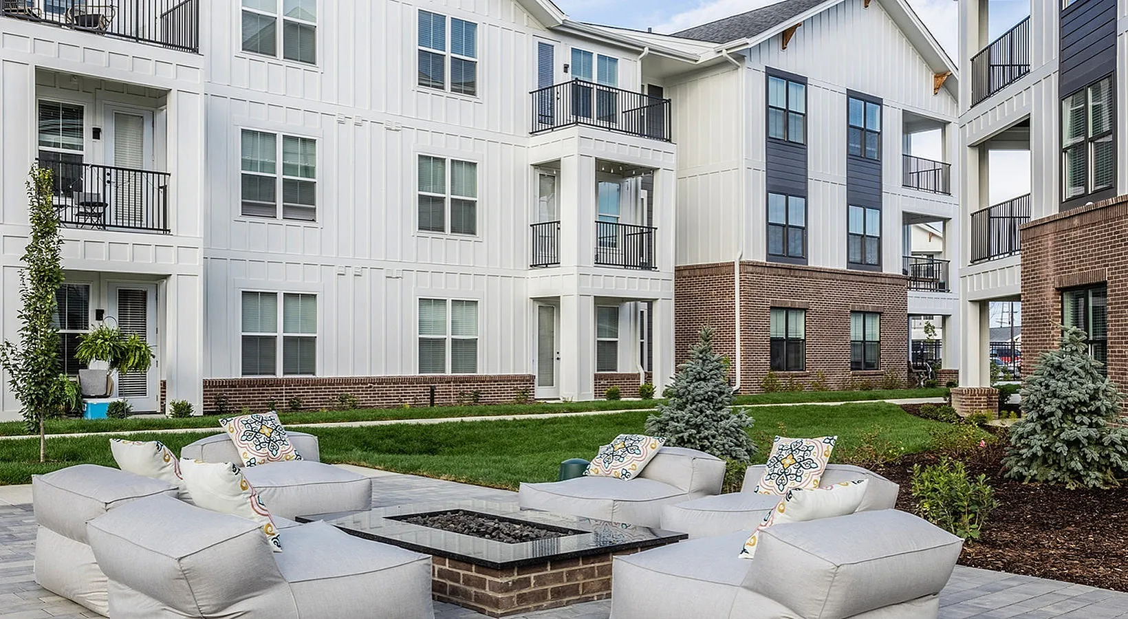 Modern apartment complex with stylish white and brick exterior, featuring spacious balconies and inviting outdoor lounge area surrounded by lush greenery.