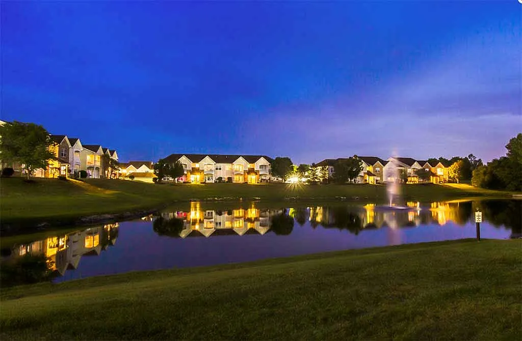 Breathtaking evening view of illuminated lakeside apartments with serene reflections and a picturesque fountain under a twilight sky.