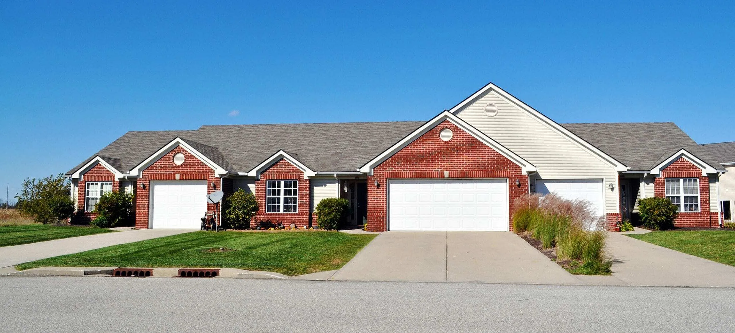 Charming duplex with classic red brick facade, manicured lawn, and double garages under a clear blue sky.