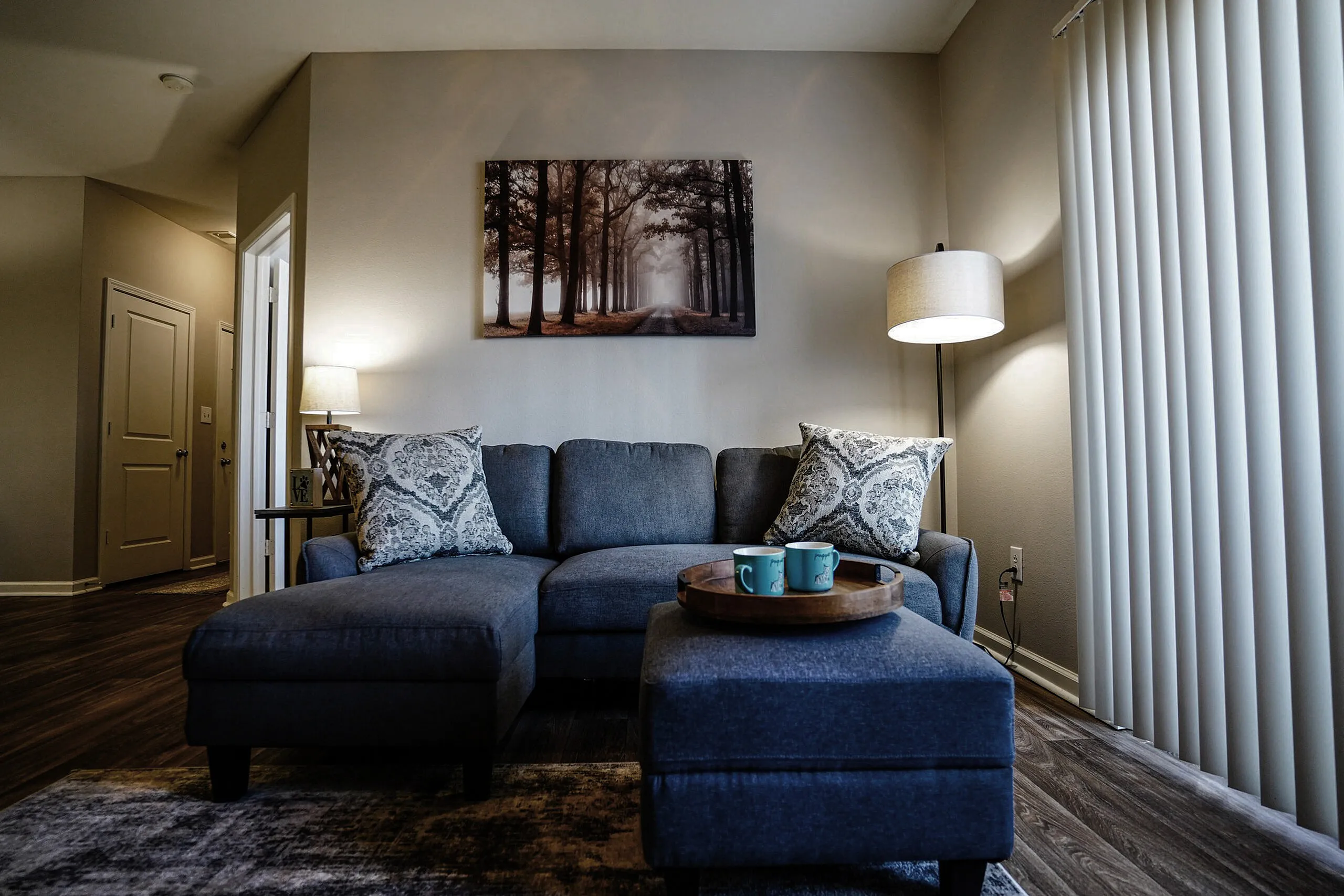 Cozy living room with a stylish gray sectional sofa, accent pillows, and warm ambient lighting. Complemented by a serene forest artwork and elegant decor, perfect for relaxation.