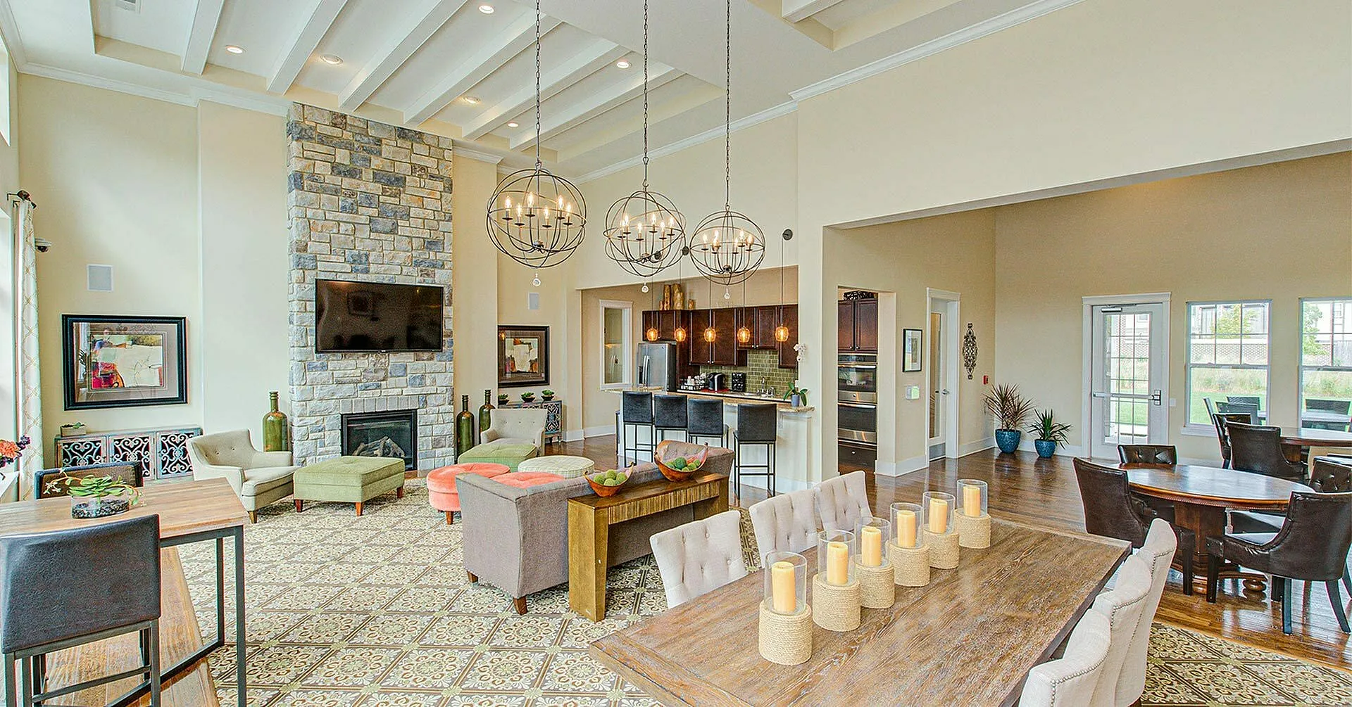Inviting open-concept living space with elegant chandeliers, stone fireplace, and modern kitchen in the background.