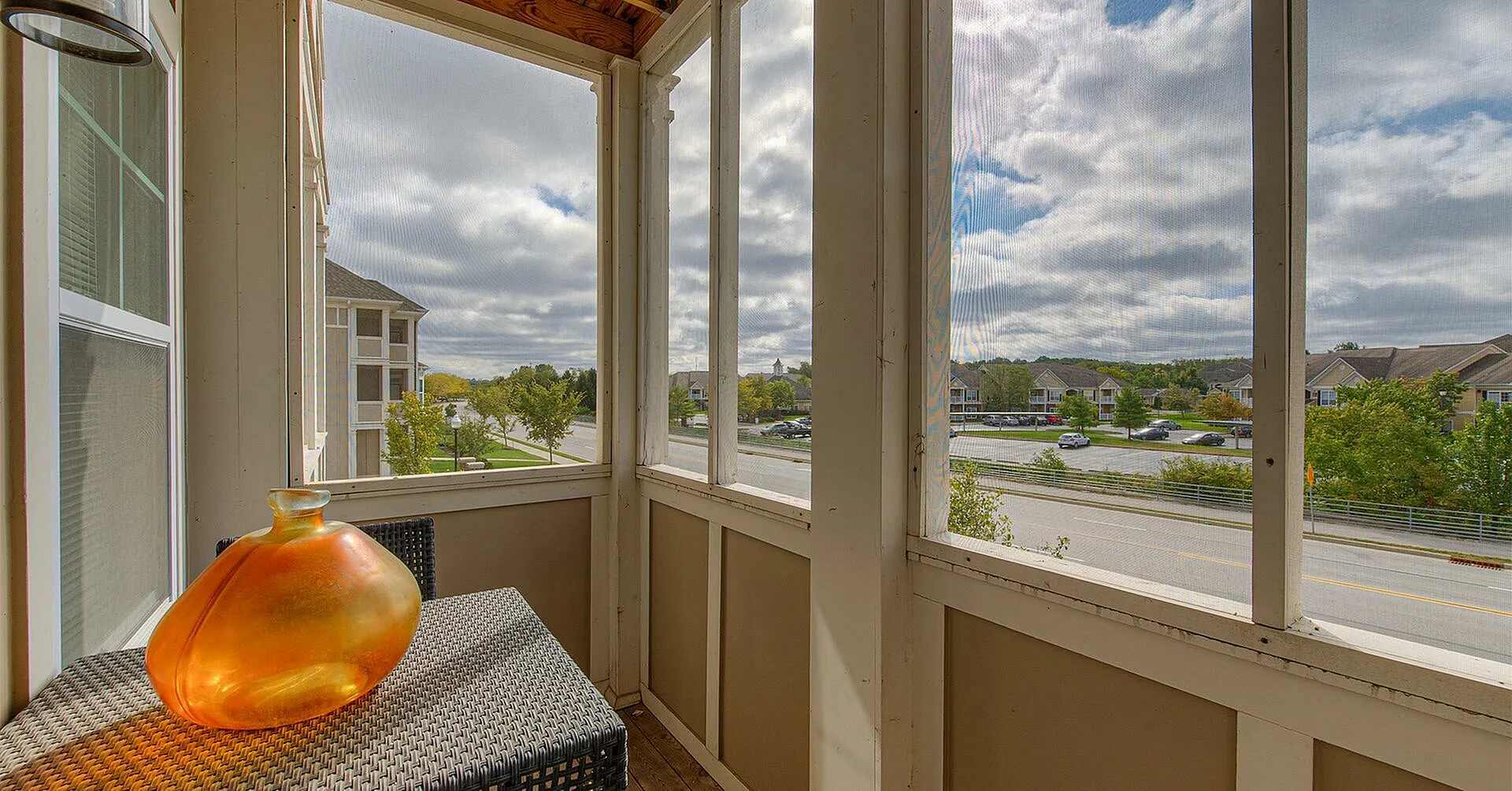 Charming enclosed balcony overlooking picturesque neighborhood views, featuring a chic wicker table with a vibrant orange vase, perfect for relaxation and enjoying the serene environment.