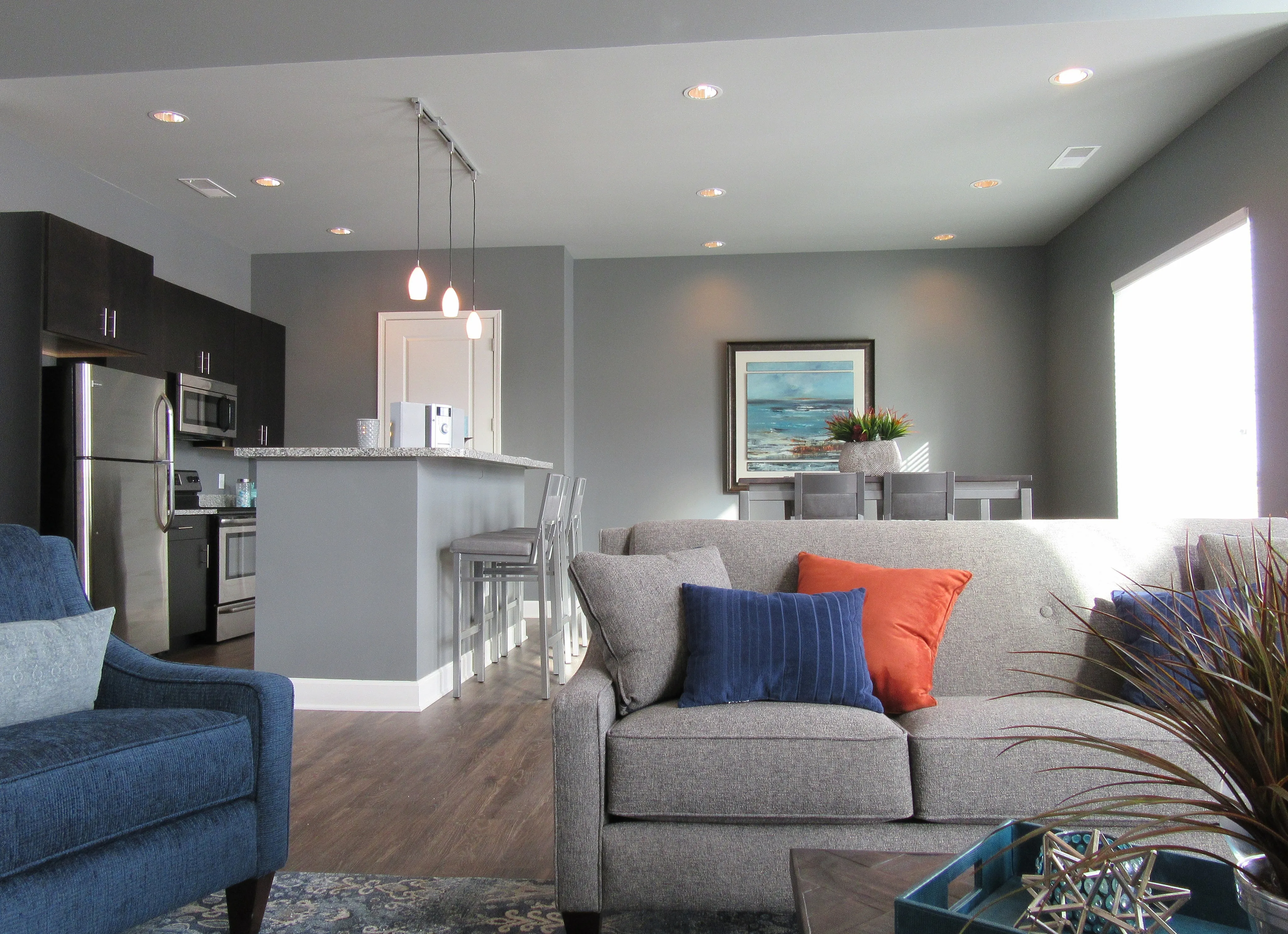 Modern open-concept living area with cozy gray sofas, accented blue and orange pillows, a stylish breakfast bar, sleek stainless steel appliances, and elegant lighting creating a warm atmosphere.