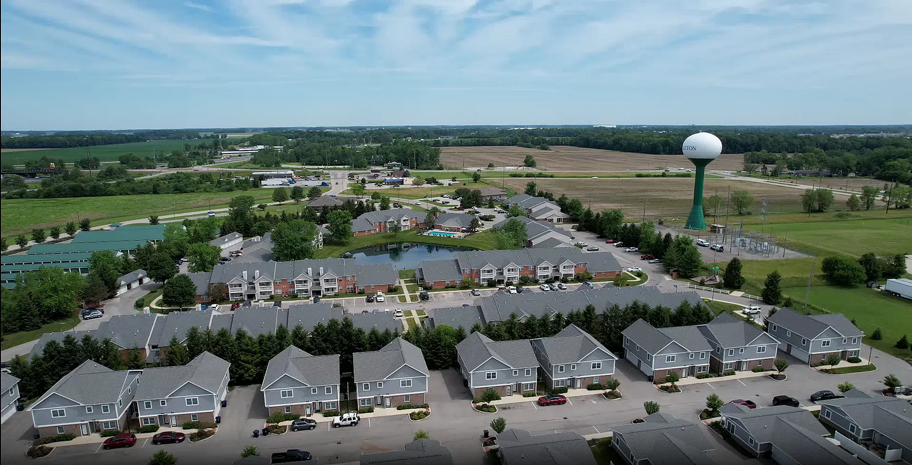 Aerial view of a picturesque apartment complex surrounded by lush greenery and open fields, featuring elegant modern townhomes and a serene pond.