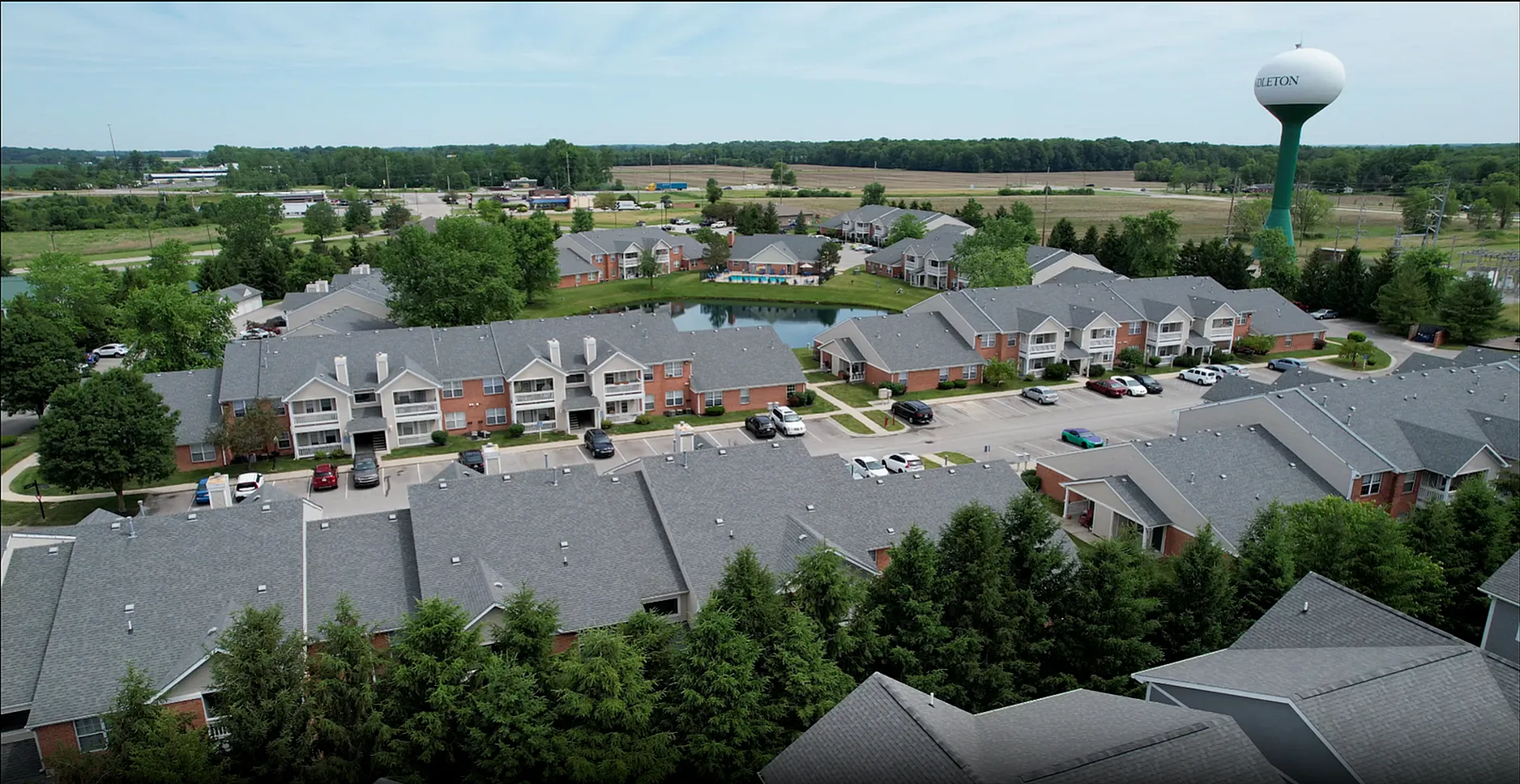 Lush aerial view of a charming apartment complex with scenic landscaping and a serene pond at its center, surrounded by verdant trees and ample parking space.